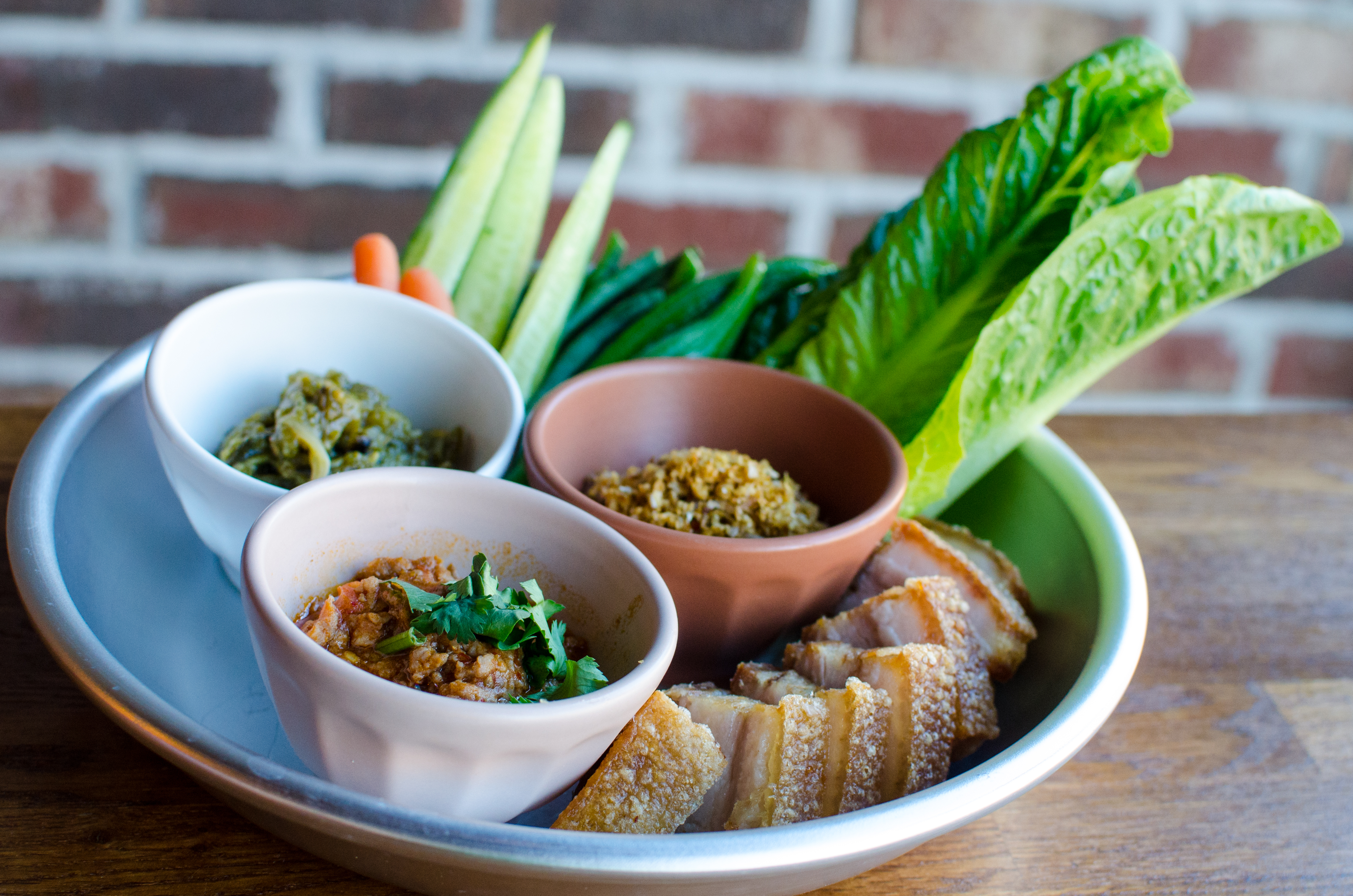 A round silver tray sits on a wooden table in front of a brick wall. On the tray, there are three small bowls — one white, one pink, one red —&nbsp;each holding a different Thai chile paste. Some vegetables and chunks of crispy pork belly are also on the tray.