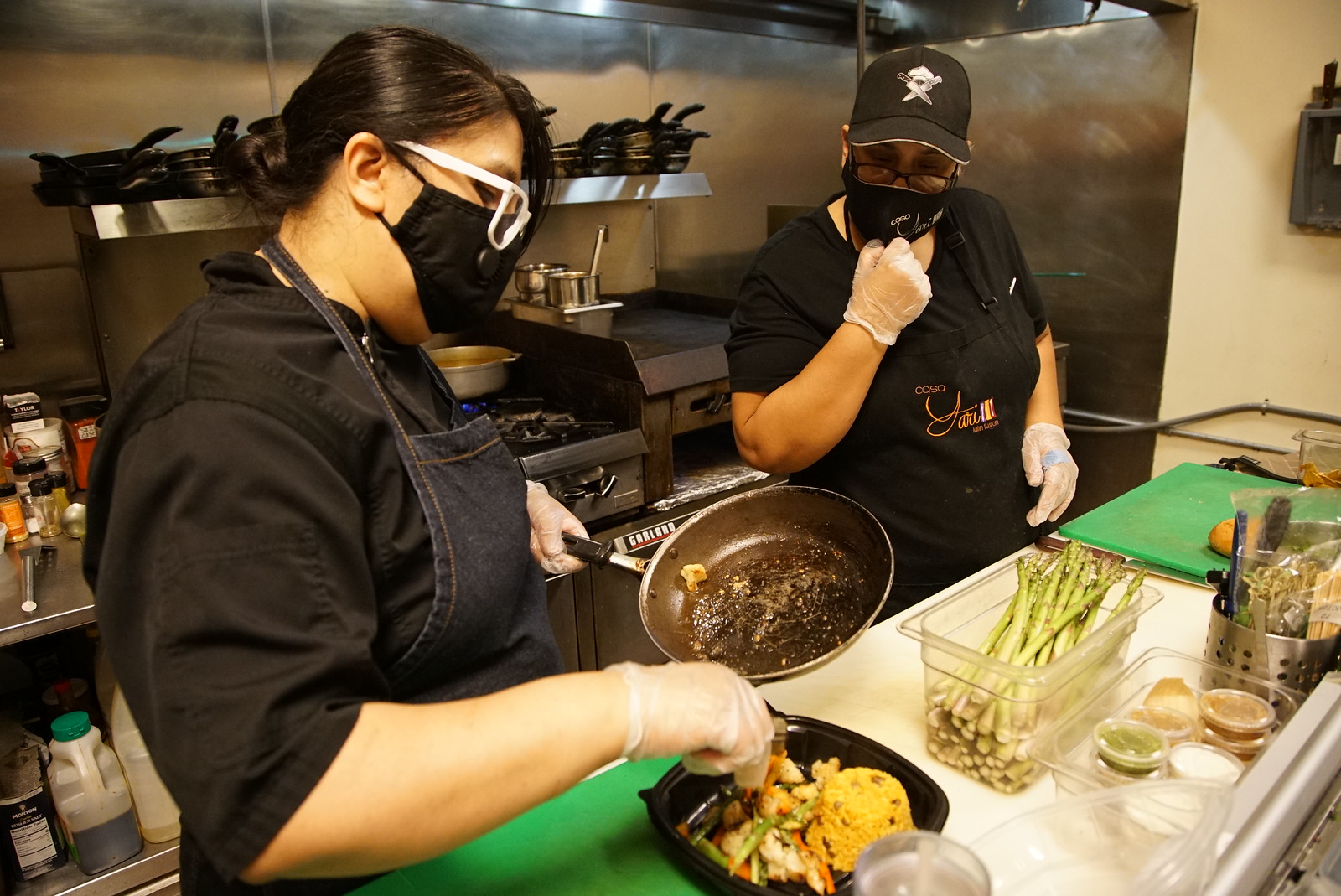 Two kitchen workers wearing masks preparing food.