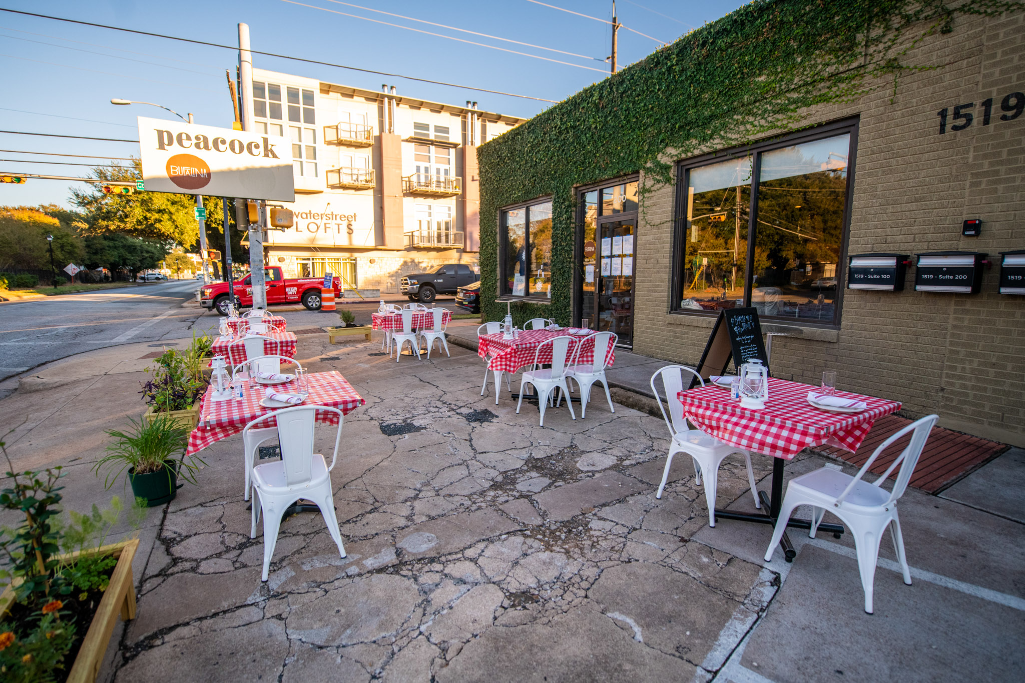 A parking lot with two-person tables with red-and-white checkered tablecloths and white chairs outside of a building covered in green ivy plants