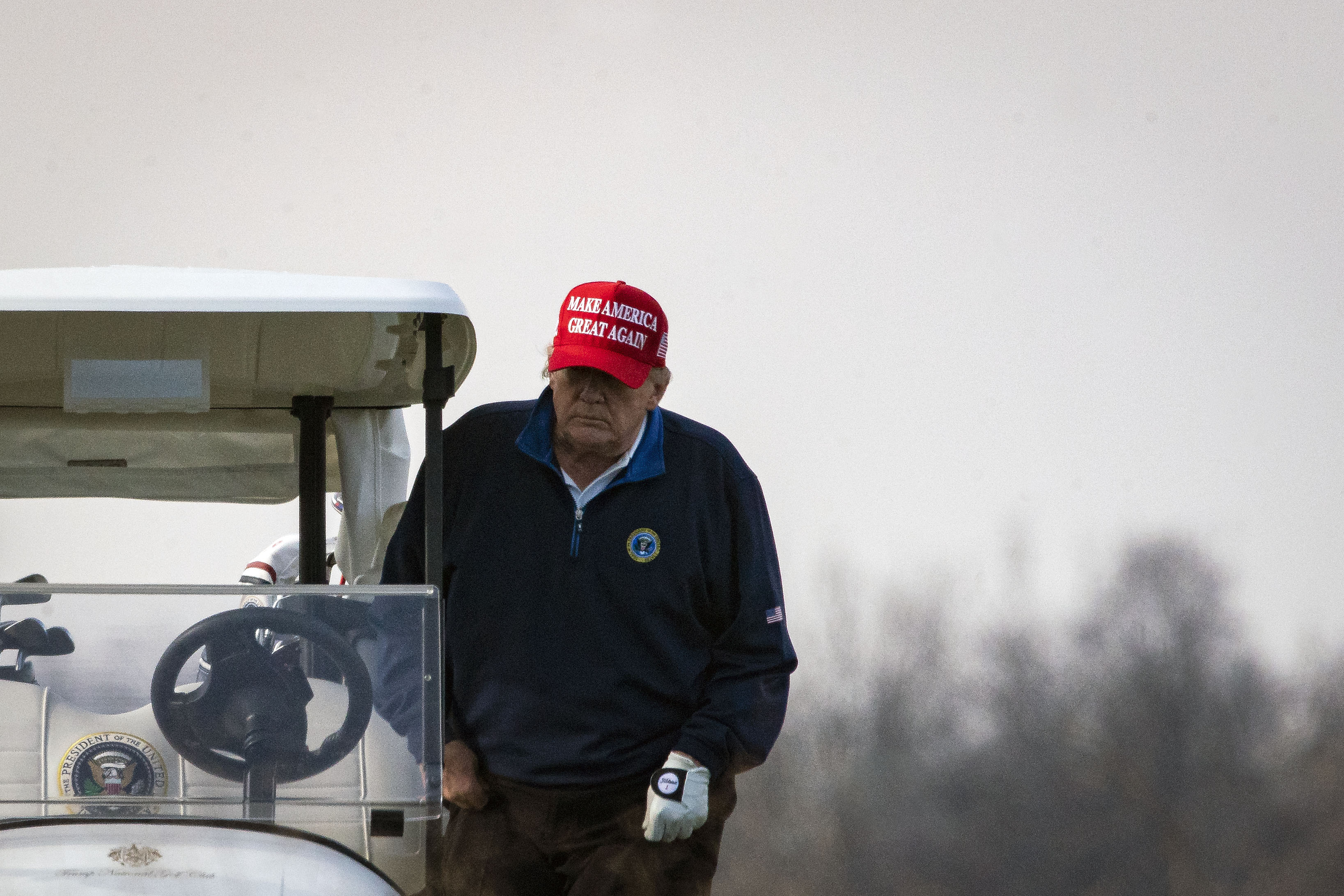 Trump, in a navy blue windbreaker featuring the seal of the president of the US and a red “Make America Great Again” baseball cap, climbs behind the wheel of a white golf cart on a cloudy day.