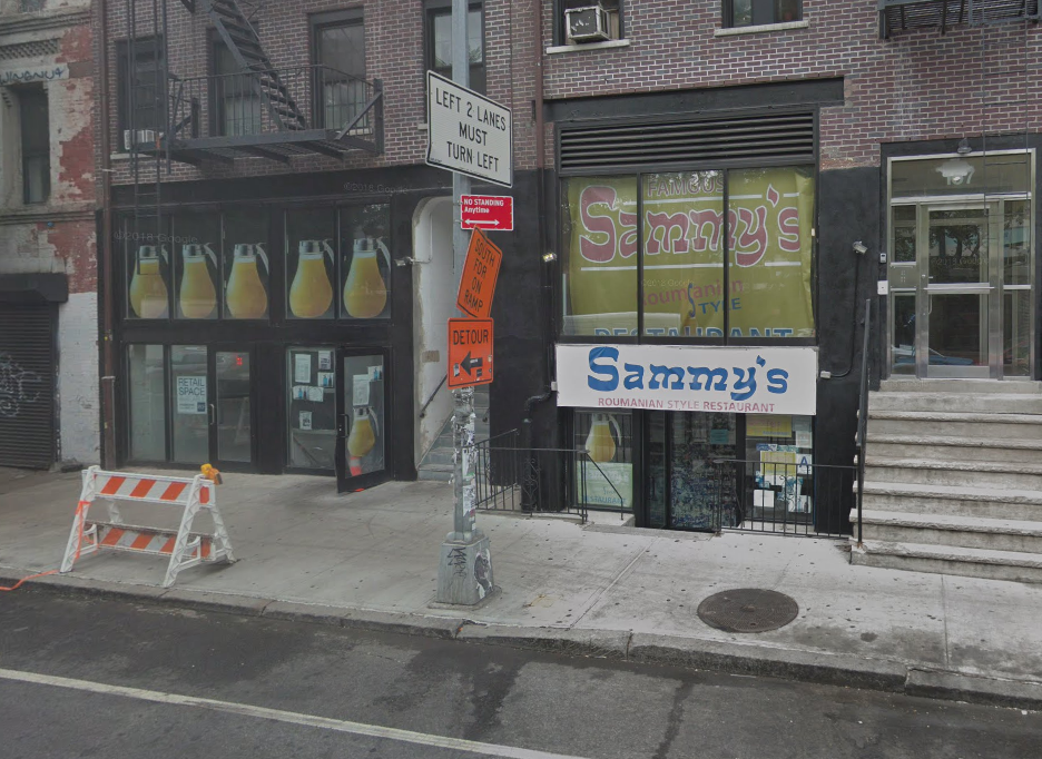 An exterior of a restaurant with a white sign and blue lettering that reads “Sammy’s” above a basement-level entry door