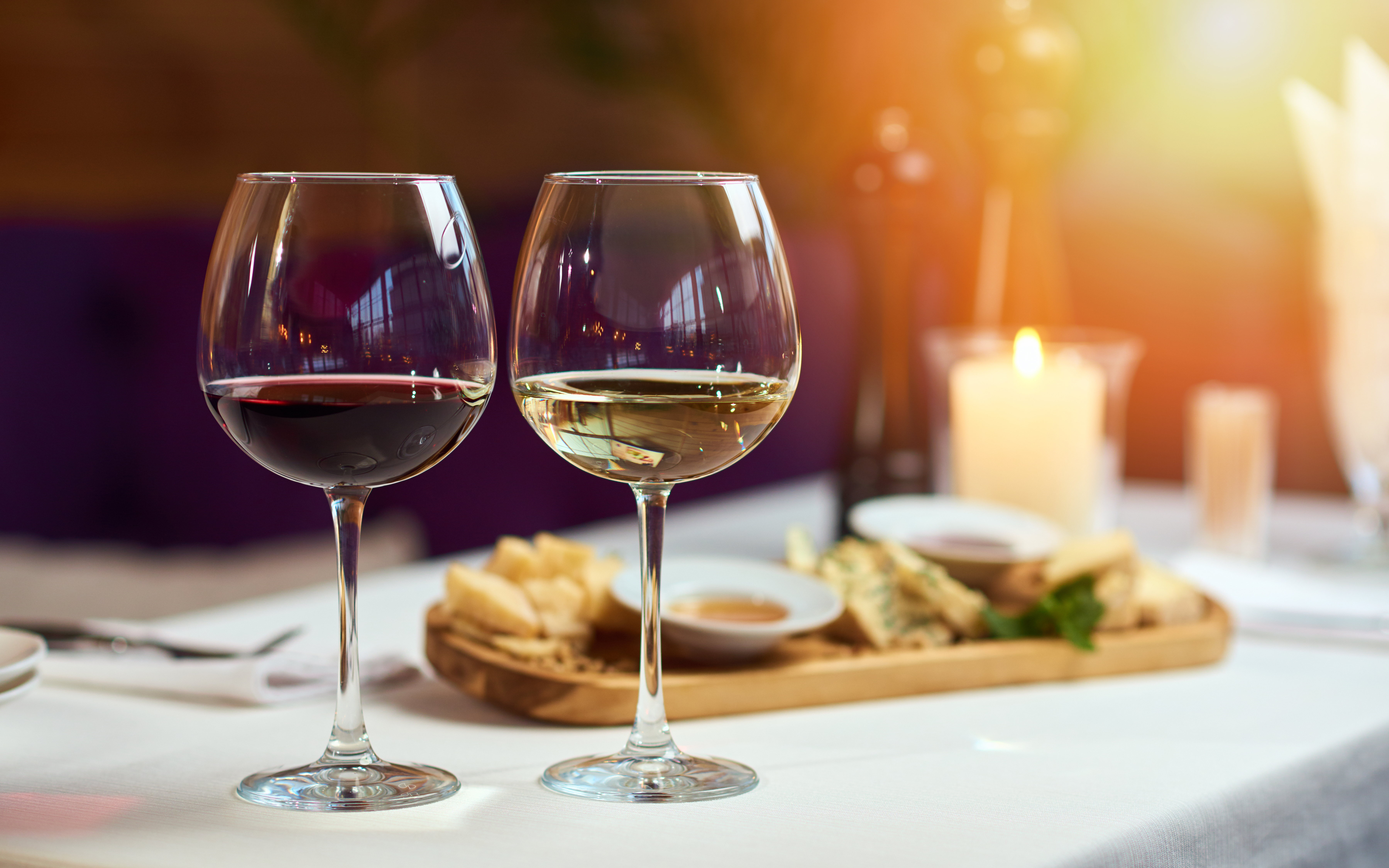 Two glasses of wine white and red standing on a table with a platter of cheeses and candle behind them in the sun light