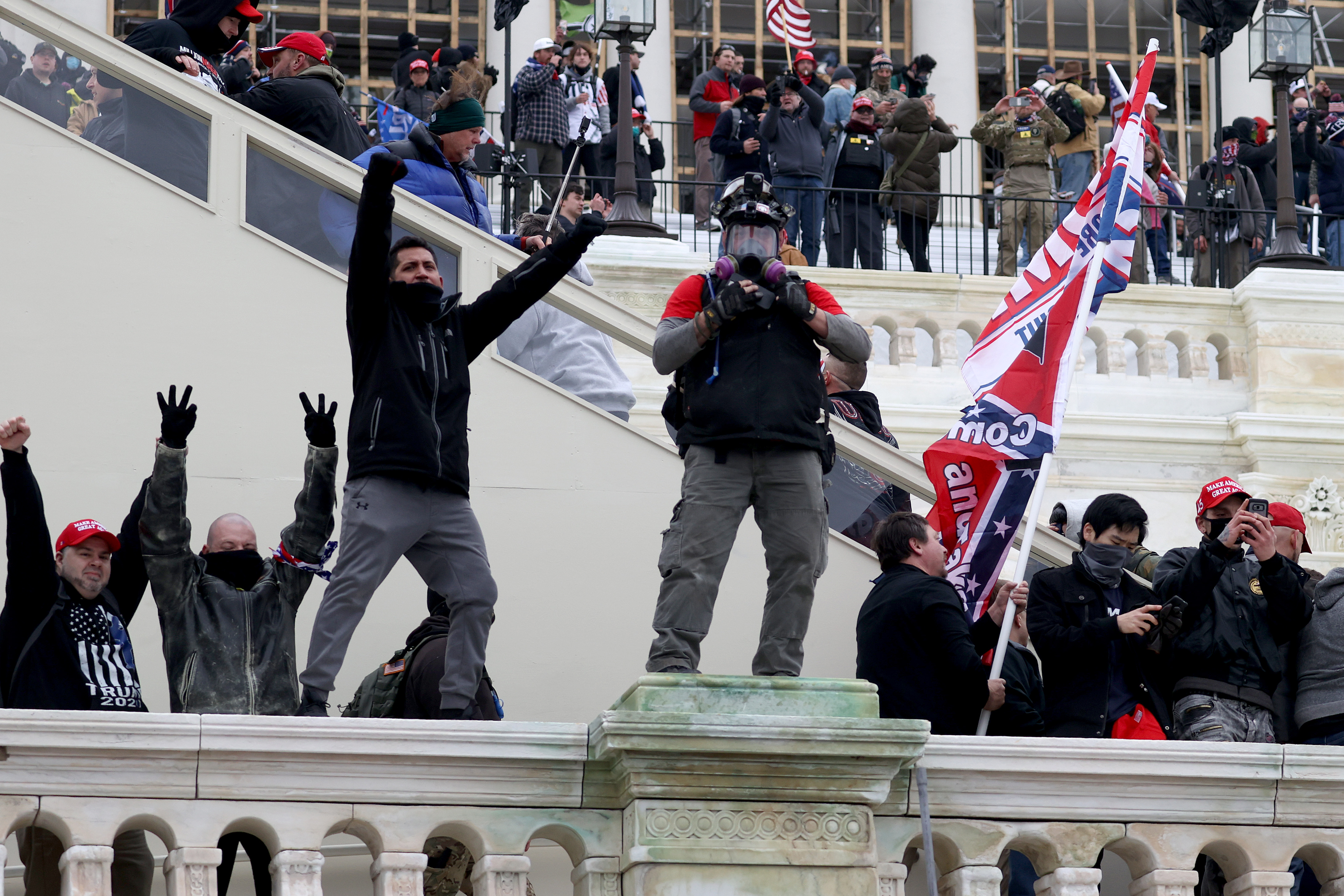 Rioters stand on the Capitol railings and stairs, holding up their arms and waving a flag.