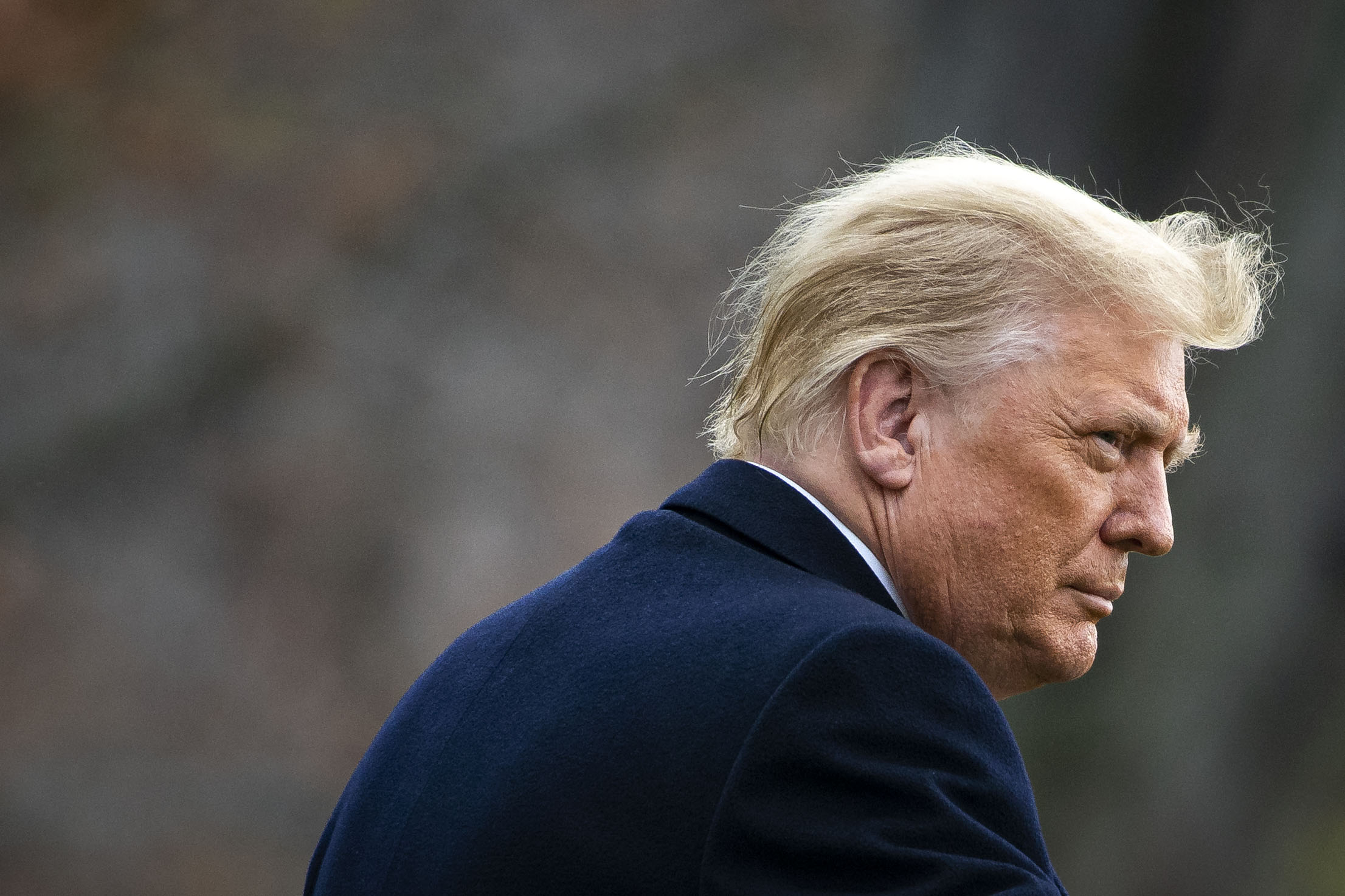 Trump, seen from the side, looks over his shoulder, frowning slightly. His white and blonde hair is being pulled back by the wind.