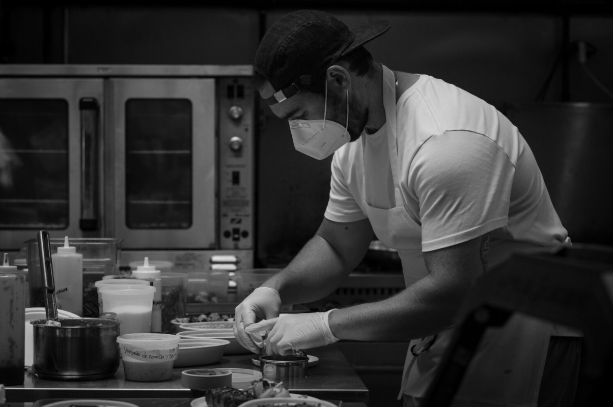 Black and white photo of chef Alejandro Tress as he prepares food in a kitchen, wearing a protective mask and a backwards cap