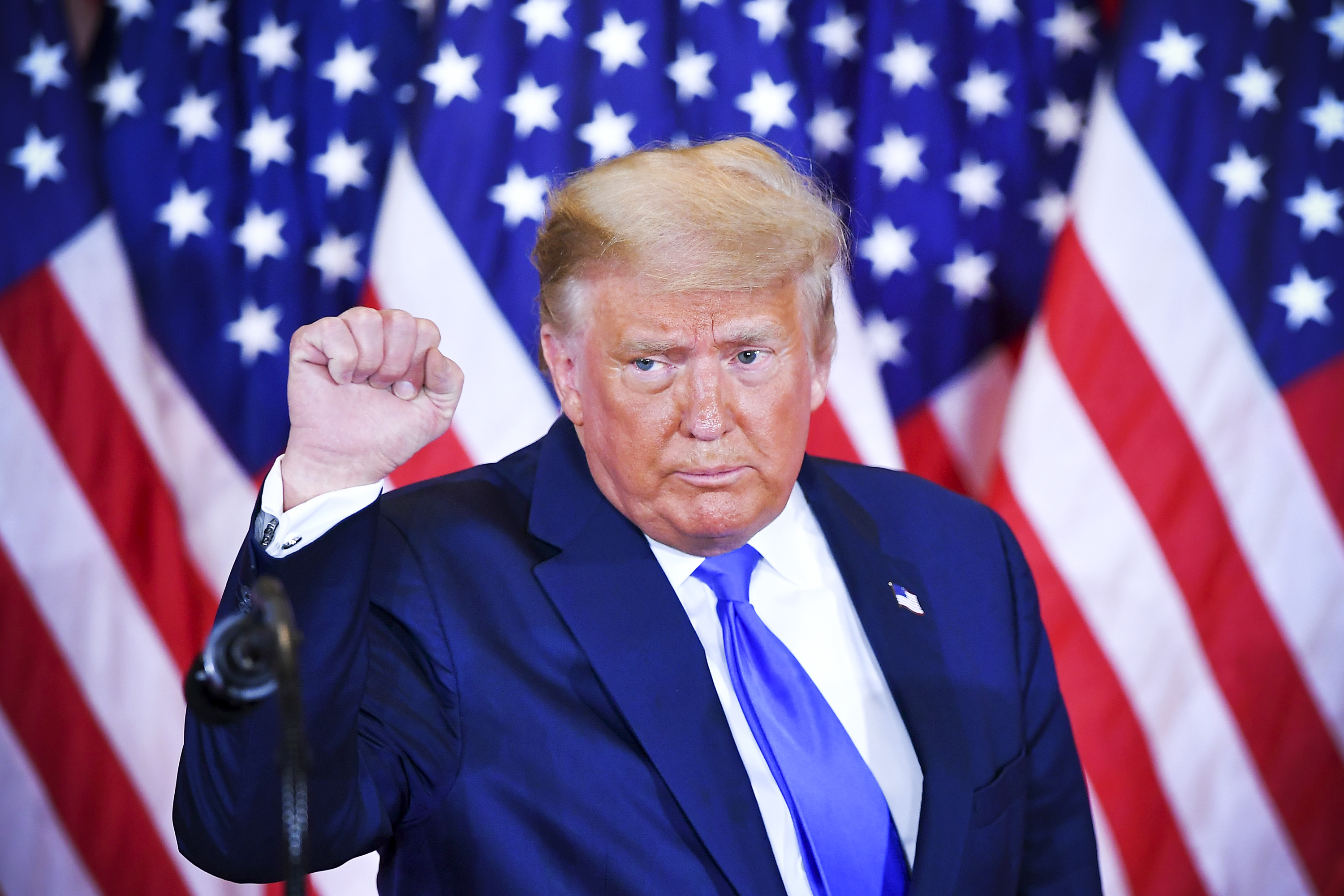 President Donald Trump, standing in front of American flags, raises his hand in a fist.