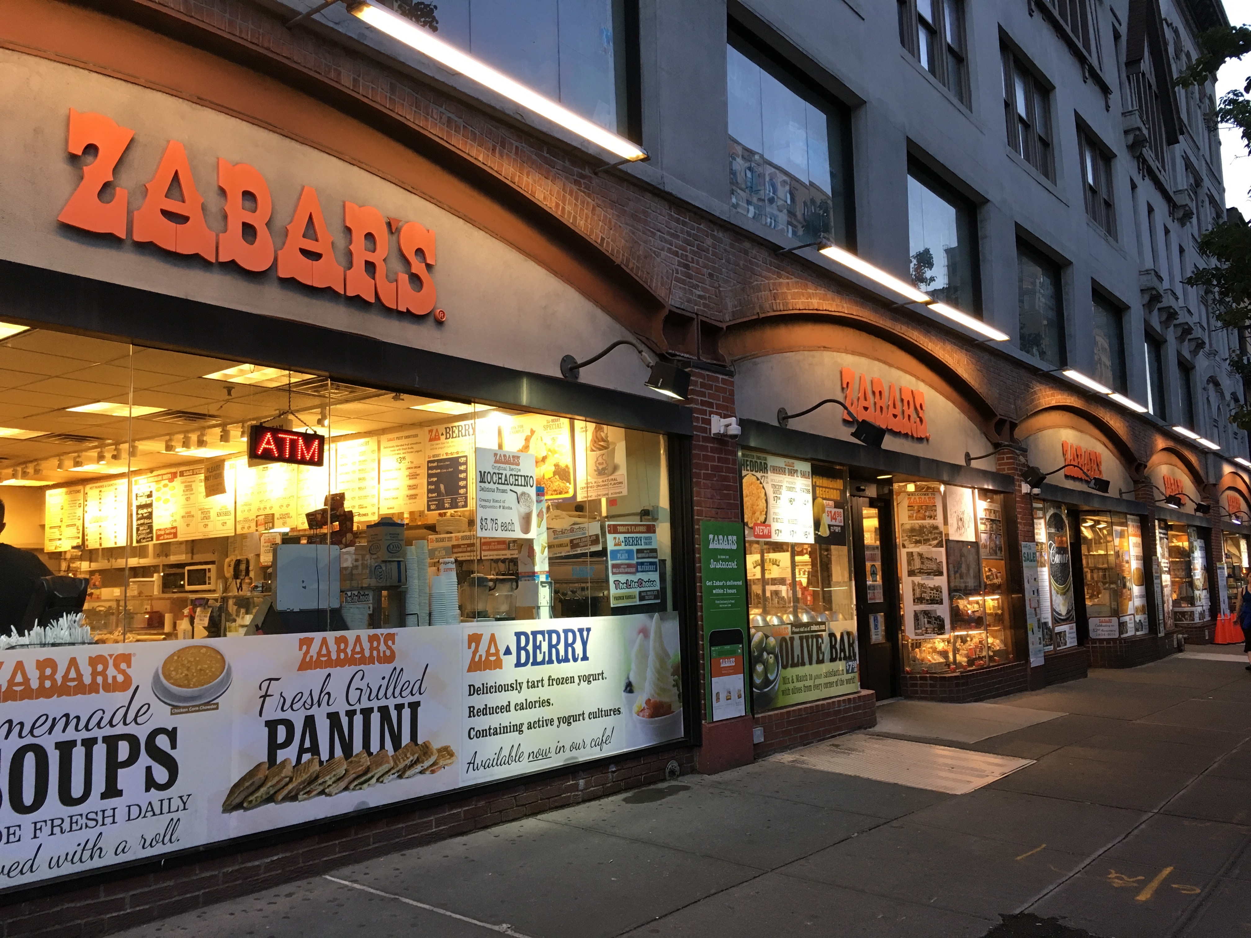 The exterior of deli and grocery store Zabar’s, orange sign illuminated, on the Upper West Side of Manhattan at dusk.