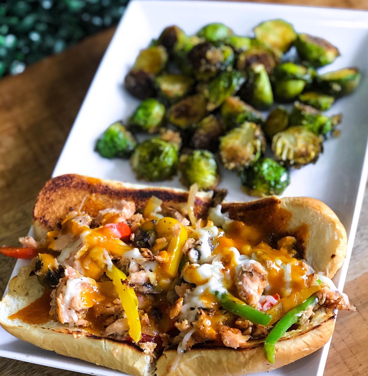 A white rectangular plate with a sandwich stuffed with grilled salmon, grilled trinity pepper mix, mushrooms, onions, and melted vegan mozzarella on brioche bread next to a side of roasted Brussels sprouts