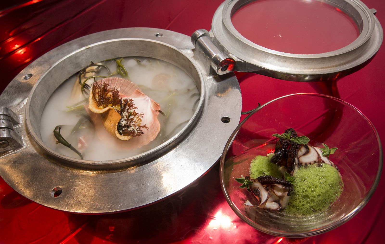 A metallic portal-shaped bowl holds a scallop dish with dry ice beside a glass bowl of octopus and green foam.