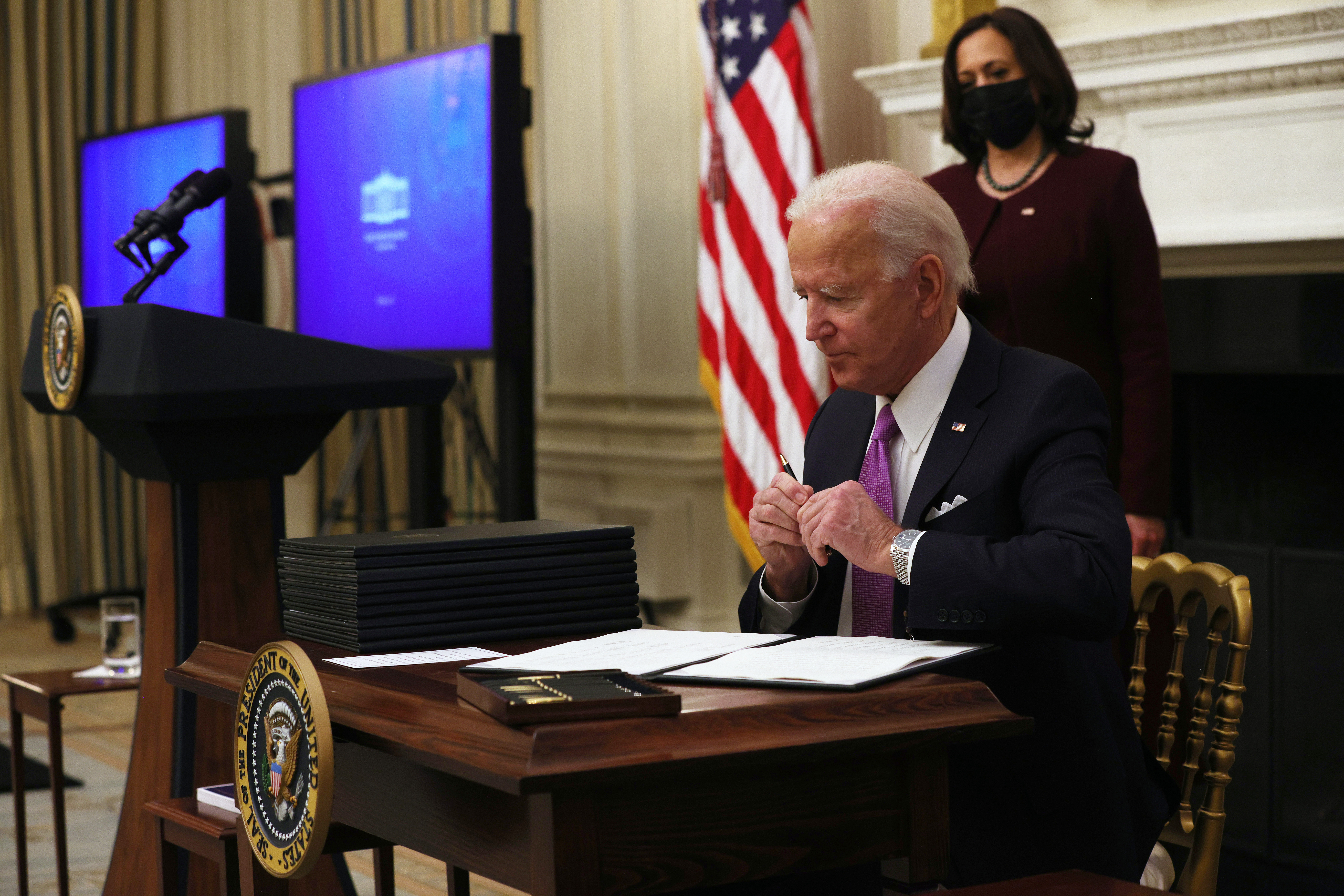 President Biden sits at a table signing documents with Vice President Kamala Harris standing behind him looking on.