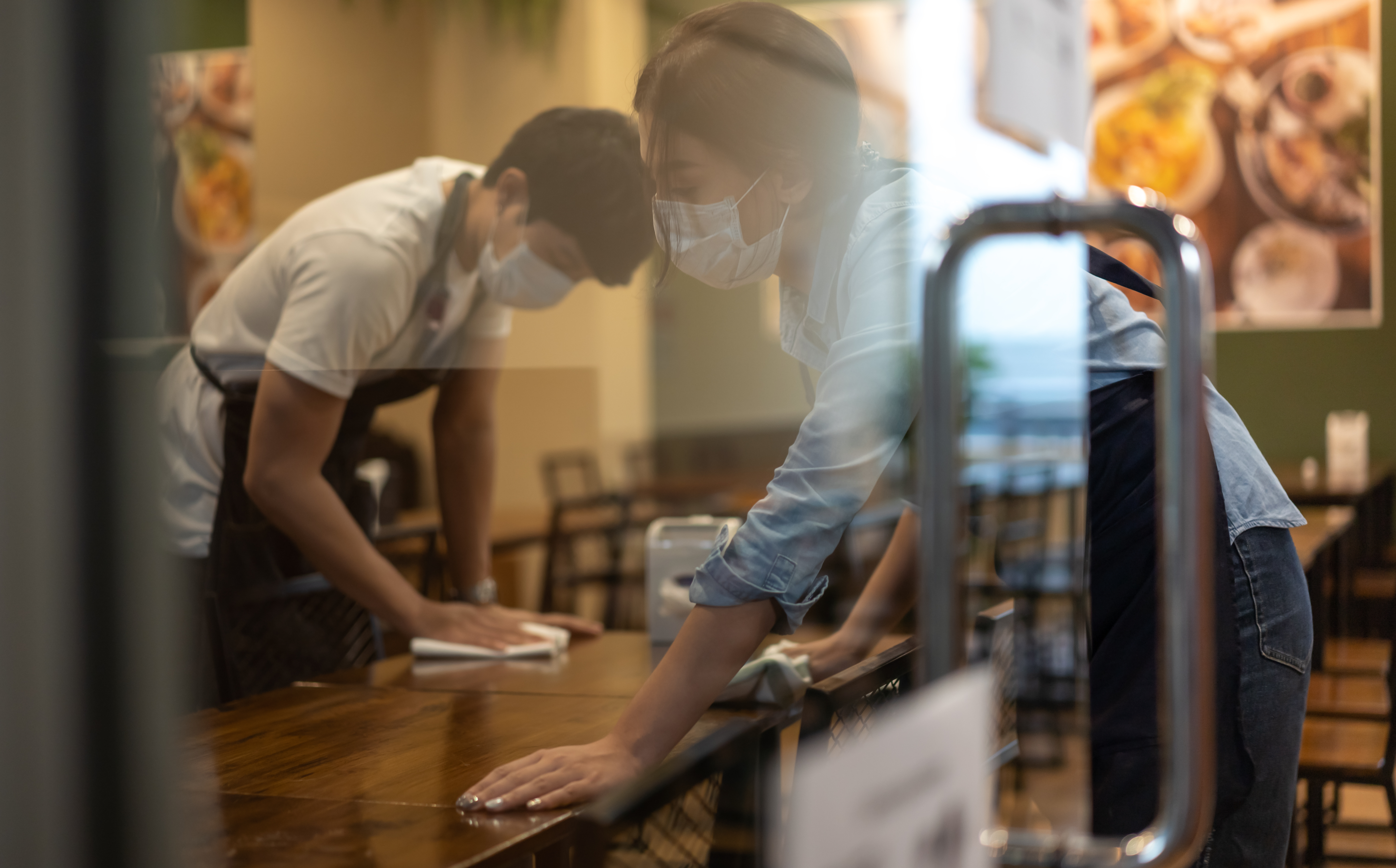 Two waiters wearing protective face mask cleans tables in a restaurant behind a window