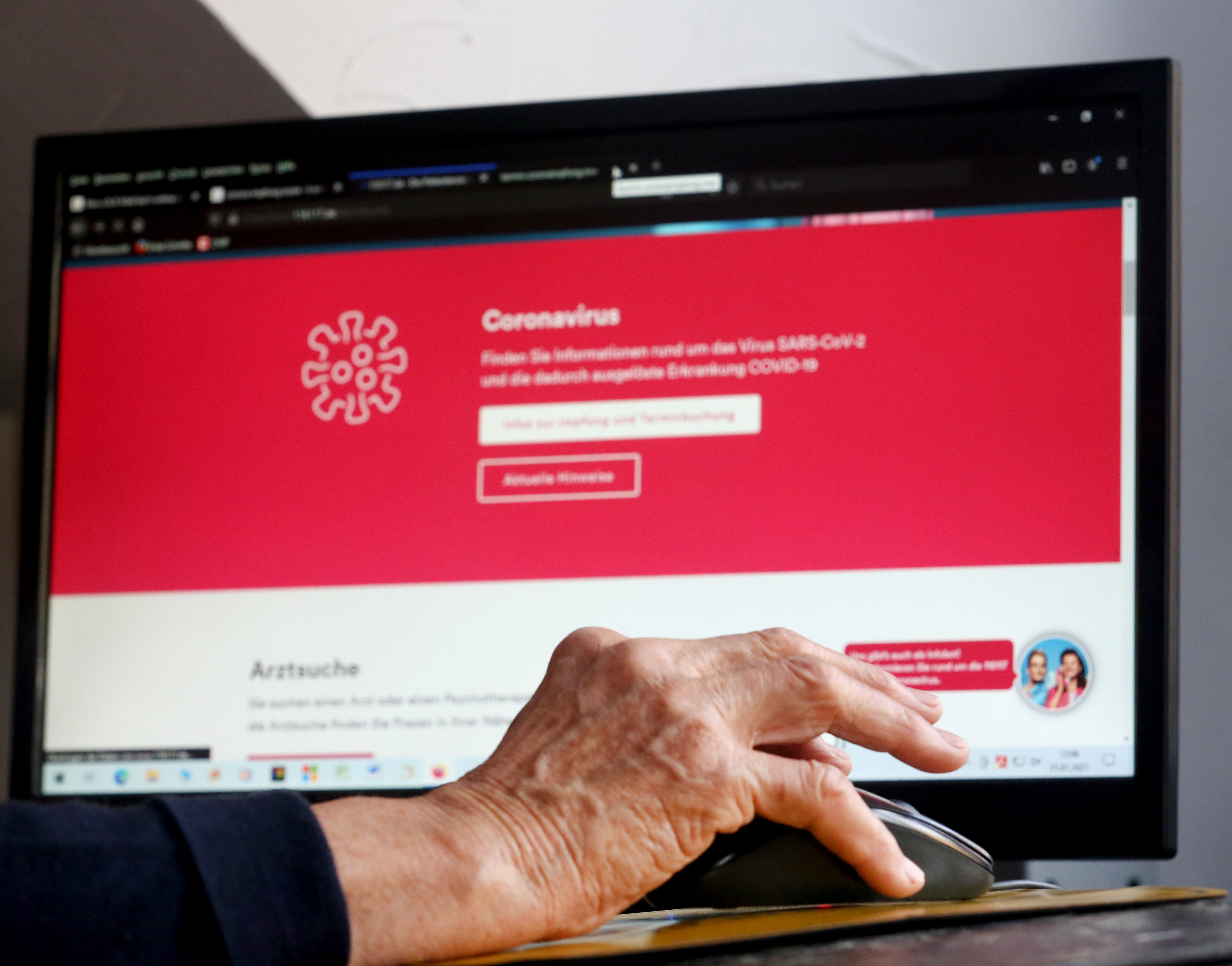 A retiree’s hand on a mouse, in front of a desktop screen showing a website about coronavirus vaccination appointments.
