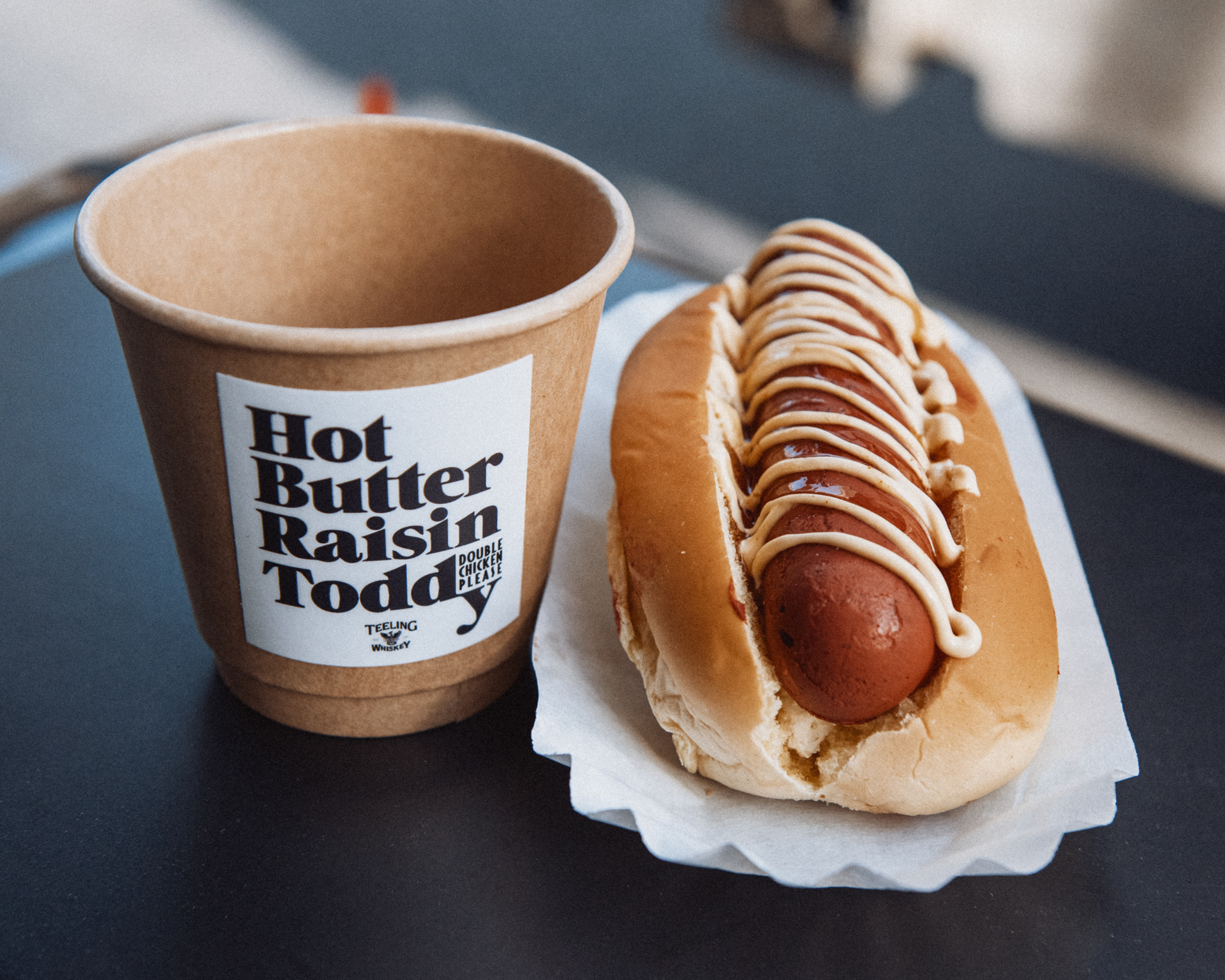 A hot dog drizzled with mustard sits next to a to-go cup that reads “Hot Butter Raisin Toddy”