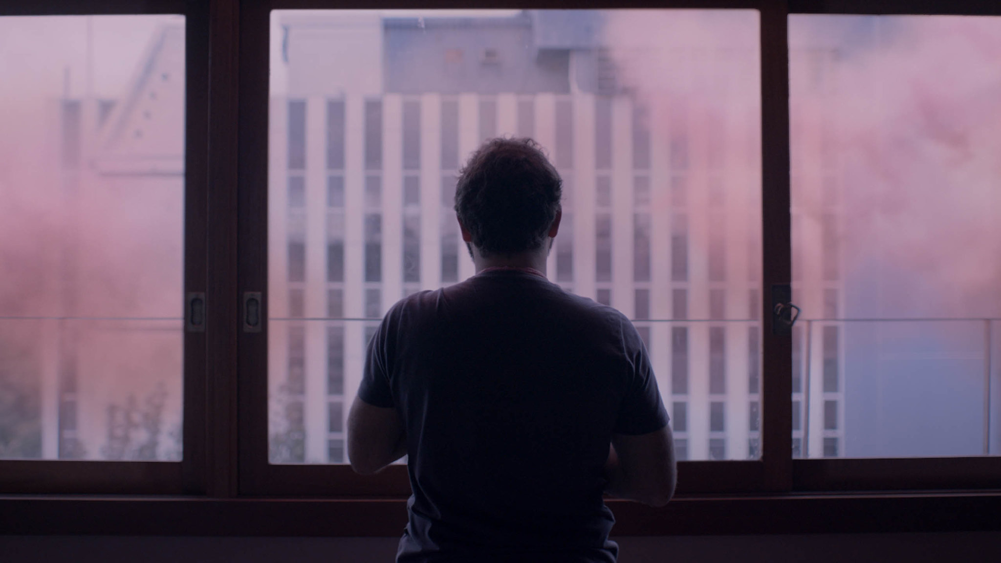 A man stands in front of a window, looking at a hazy pink cloud outside.