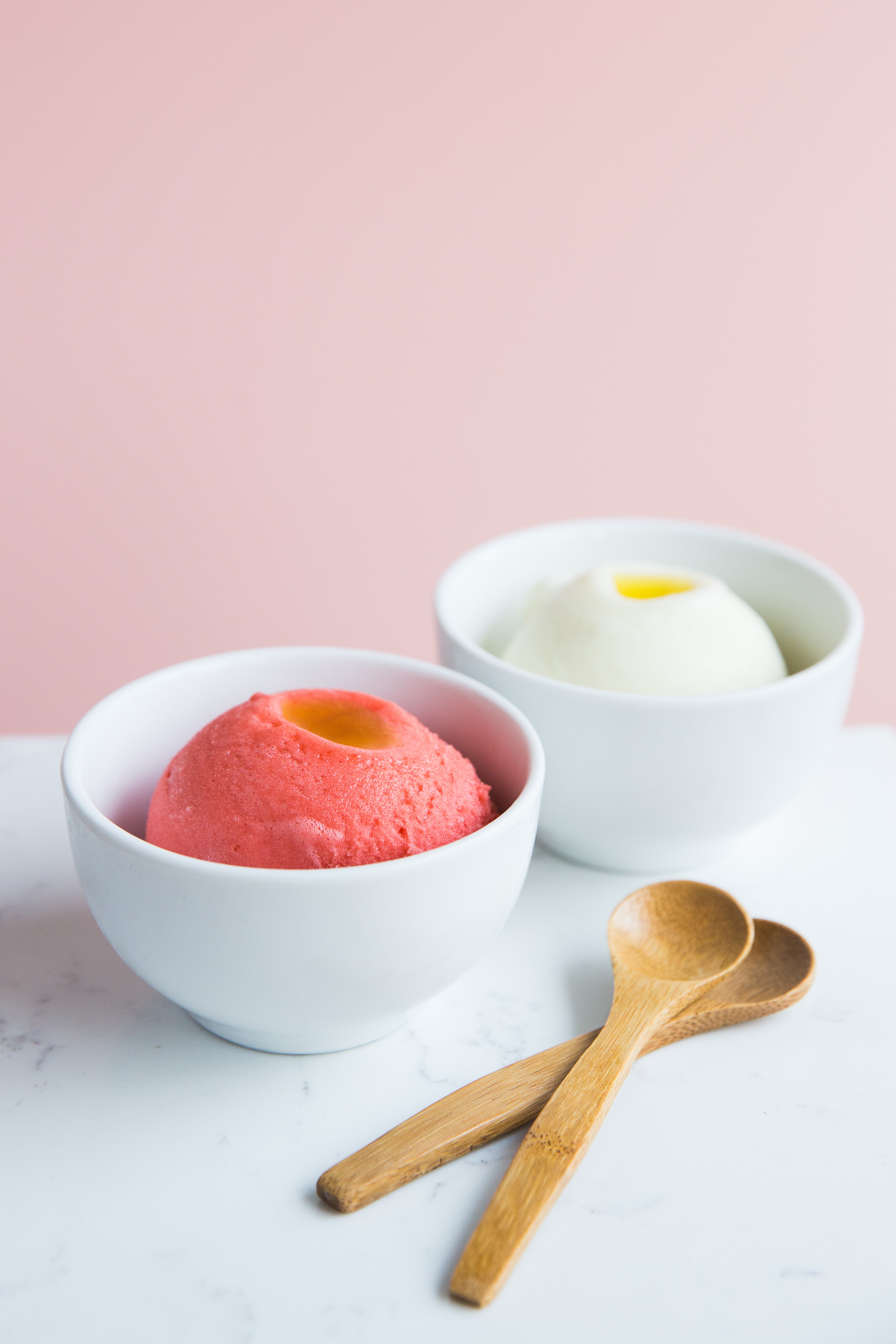 Two white cups of gelato (one scoop pink, one white), with two wooden spoons laying off to the side