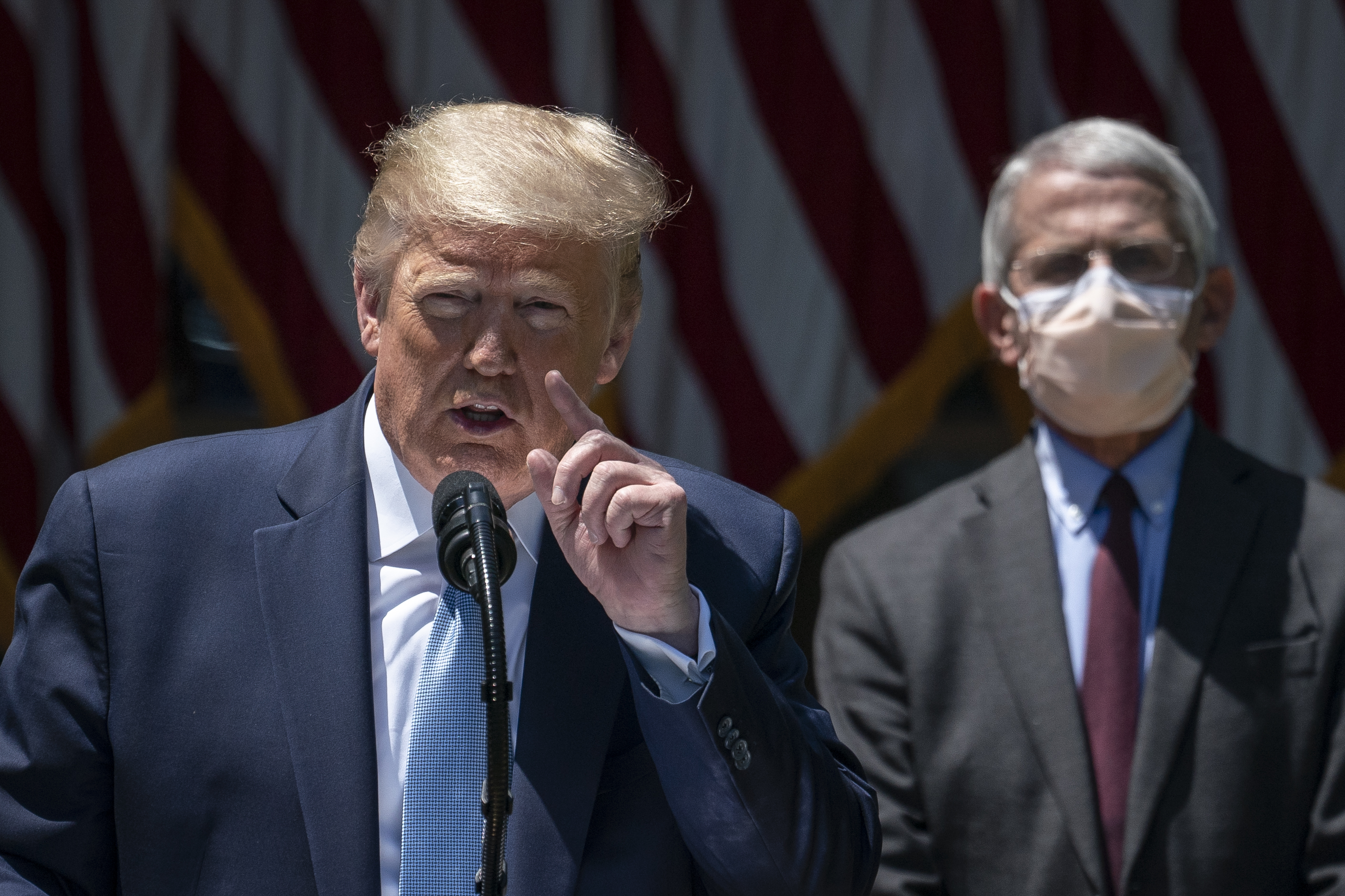 Trump, in a navy blue suit and light blue tie, speaks at a microphone, gesturing with one hand, as Fauci stands beside him, wearing a white mask, gray suit, and dark red tie.