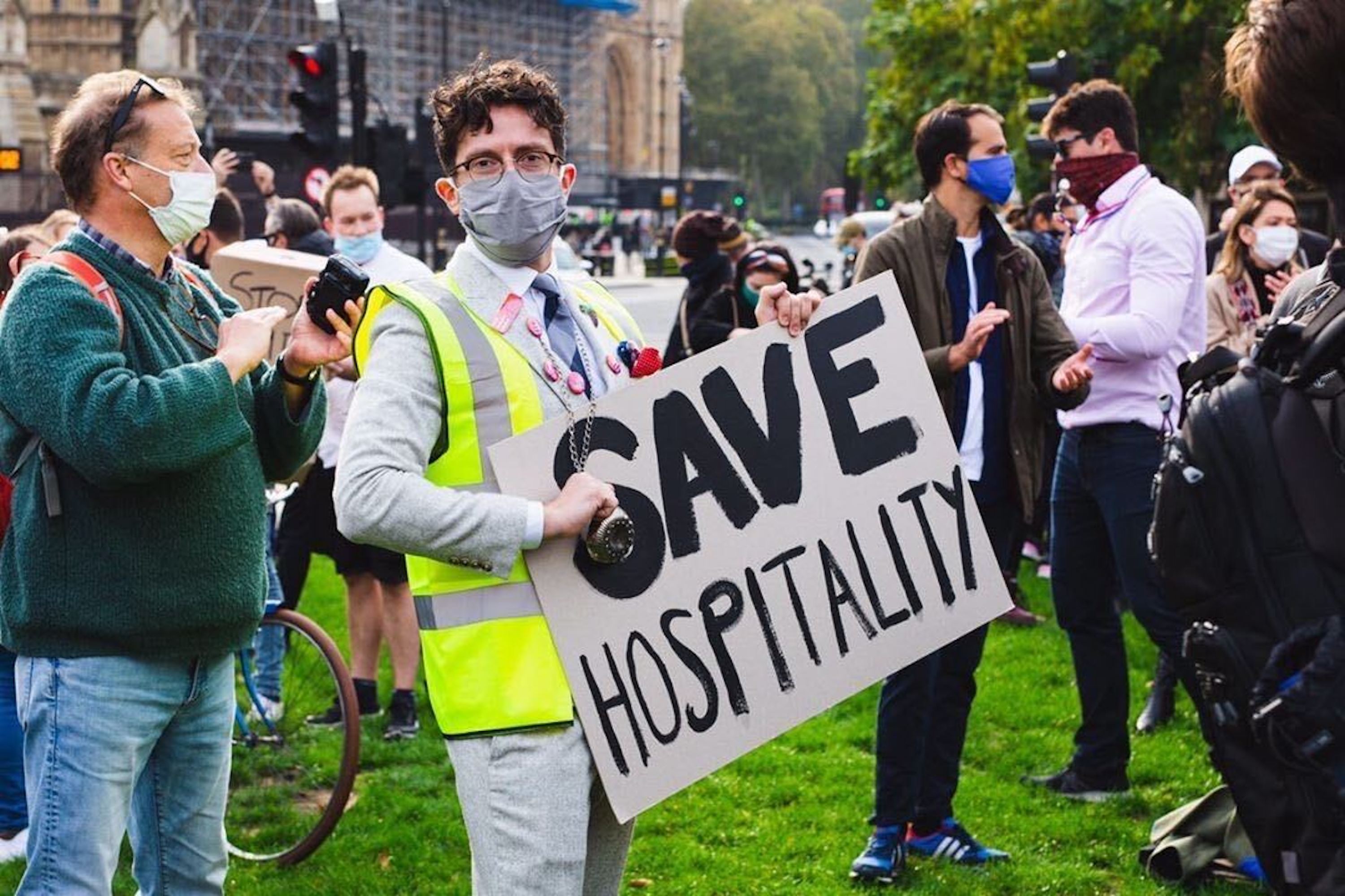 A protester wearing a mask and a hi-vis jacket holds up a placard that reads “save hospitality,” in capital letters, at Parliament Square in London. He is surrounded by fellow protesters, also in masks