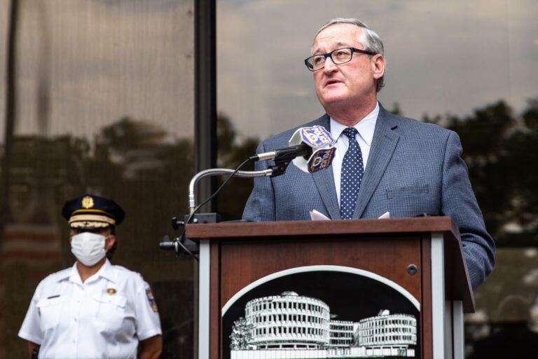 Philadelphia Mayor Jim Kenney speaks at a podium in front of glass windows, as a police officer wears a dress uniform and protective mask behind him.