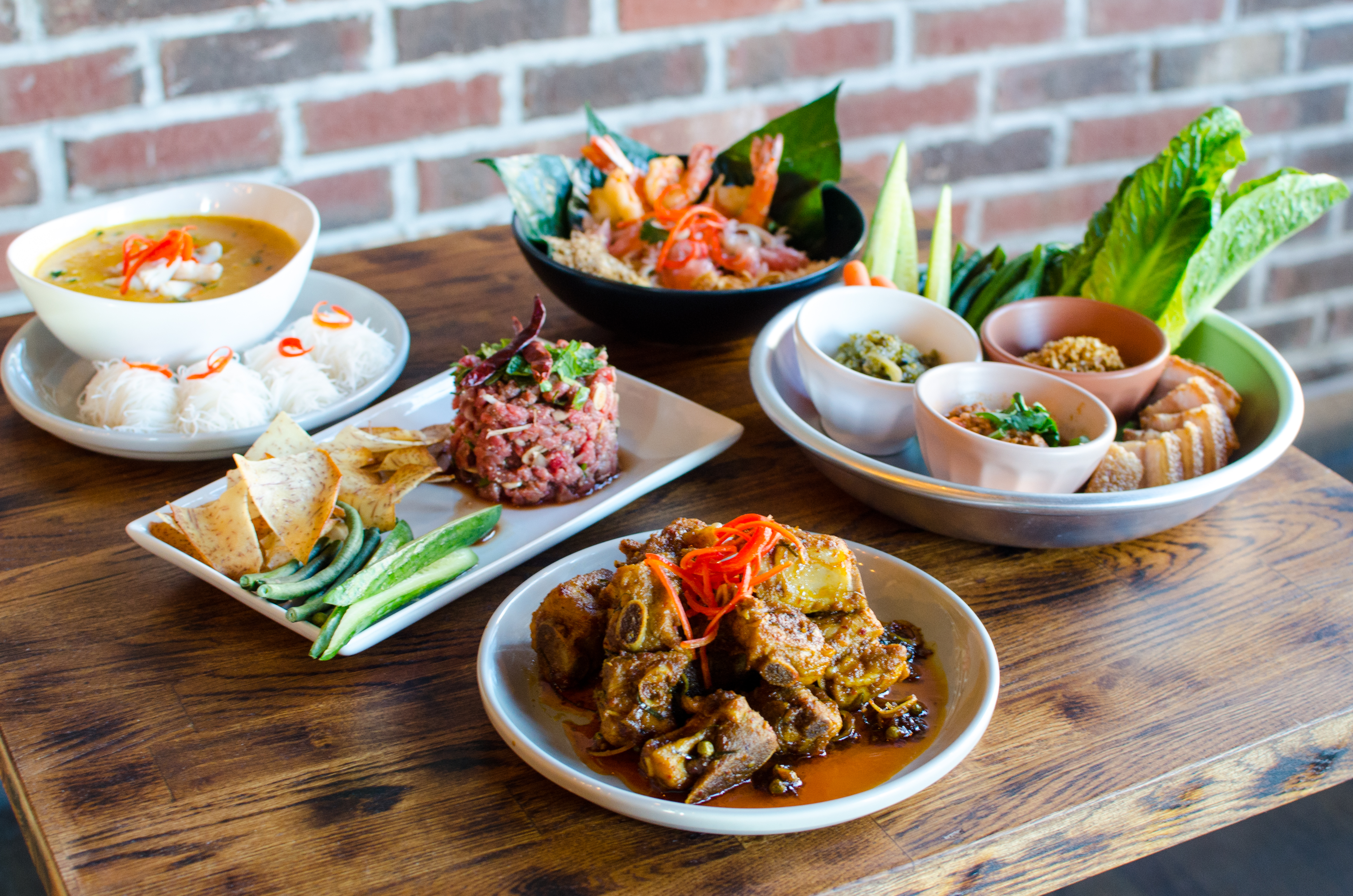 Five Thai dishes sit on a wooden table in front of a brick wall. There’s a curry, a pomelo salad with shrimp, a large pile of pork ribs, and more.