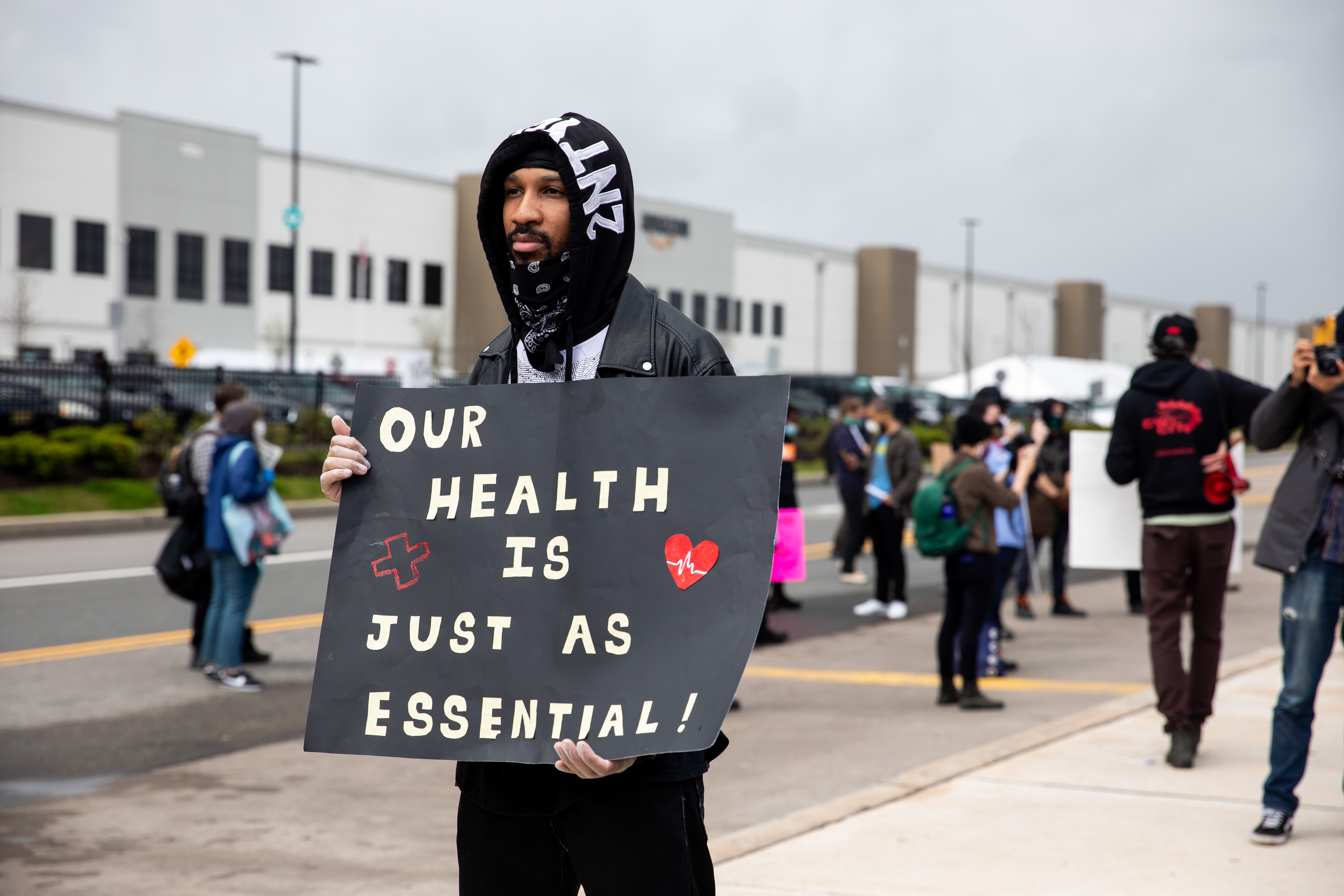A protester standing outside an Amazon facility holds a sign that reads “Our health is just as essential.”