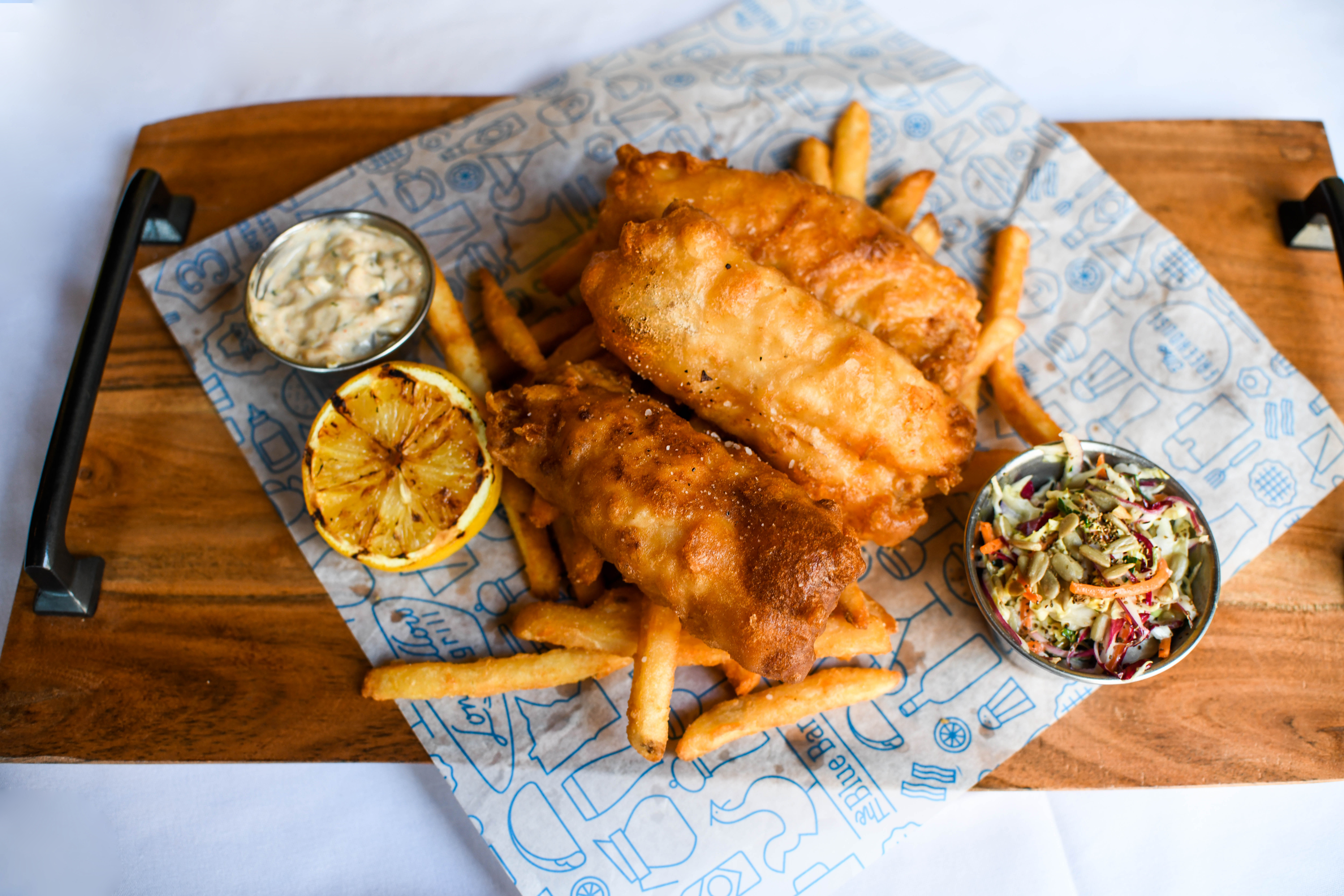 Fish and chips with lemon, coleslaw, and tartar sauce over a blue and white paper on a wooden board. 