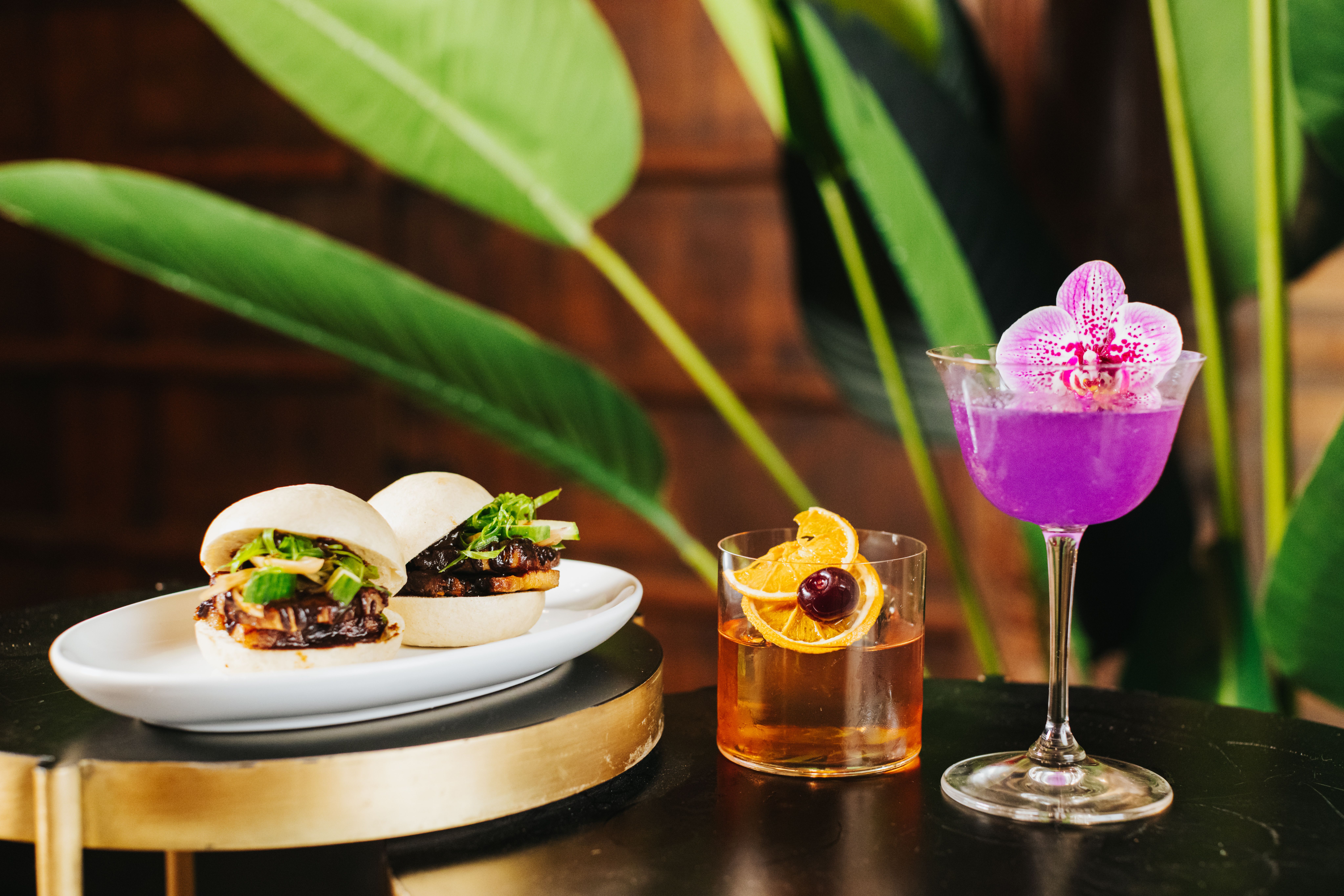 A plate of bao buns filled with pork belly next to a short brown cocktail and a tall purple cocktail garnished with an orchid.