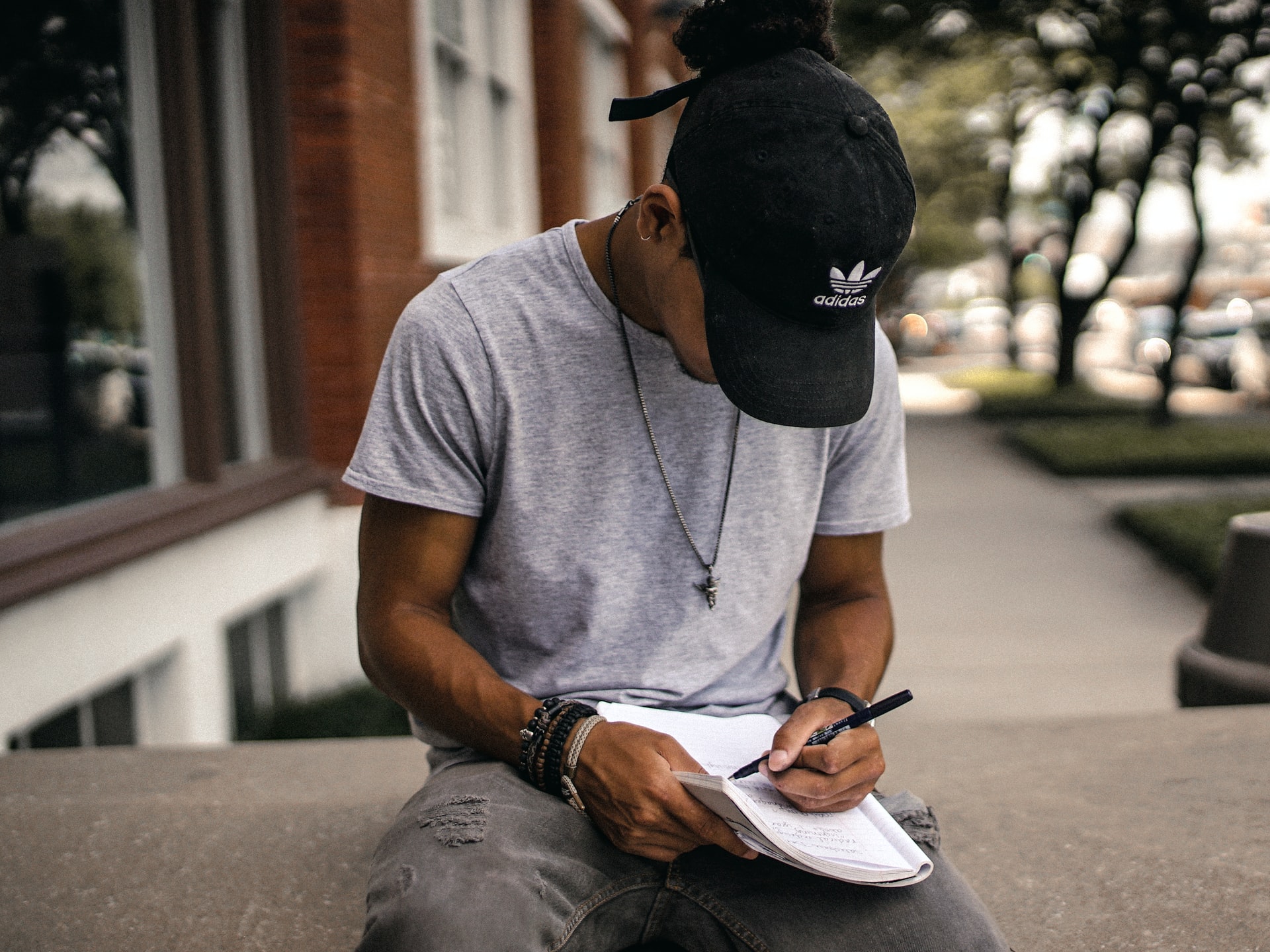 Student wearing a baseball cap sitting outside writing in a notebook.