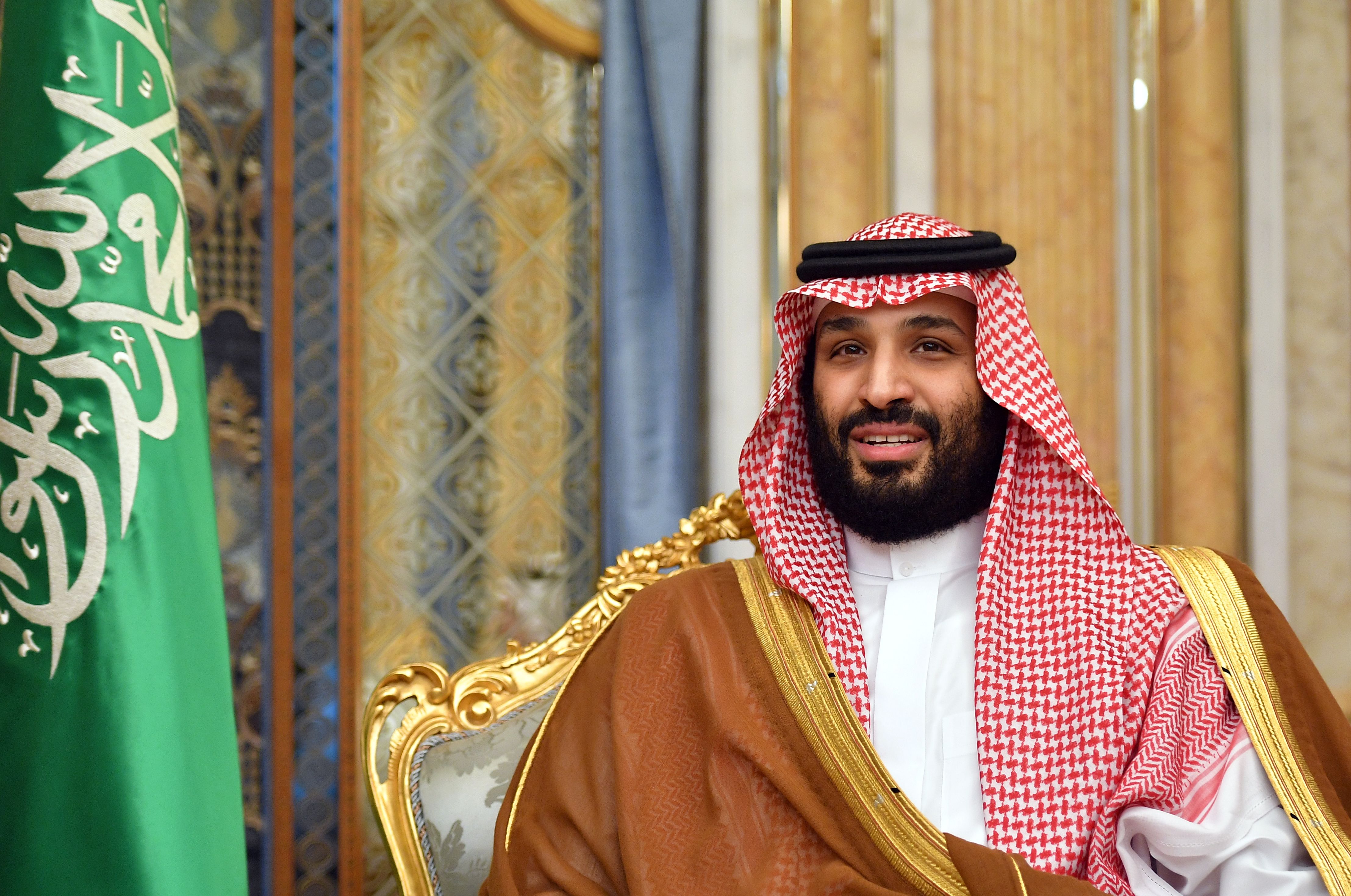 The crown prince, in traditional headscarf and robes, sits in a gold-backed chair with a Saudi flag nearby.