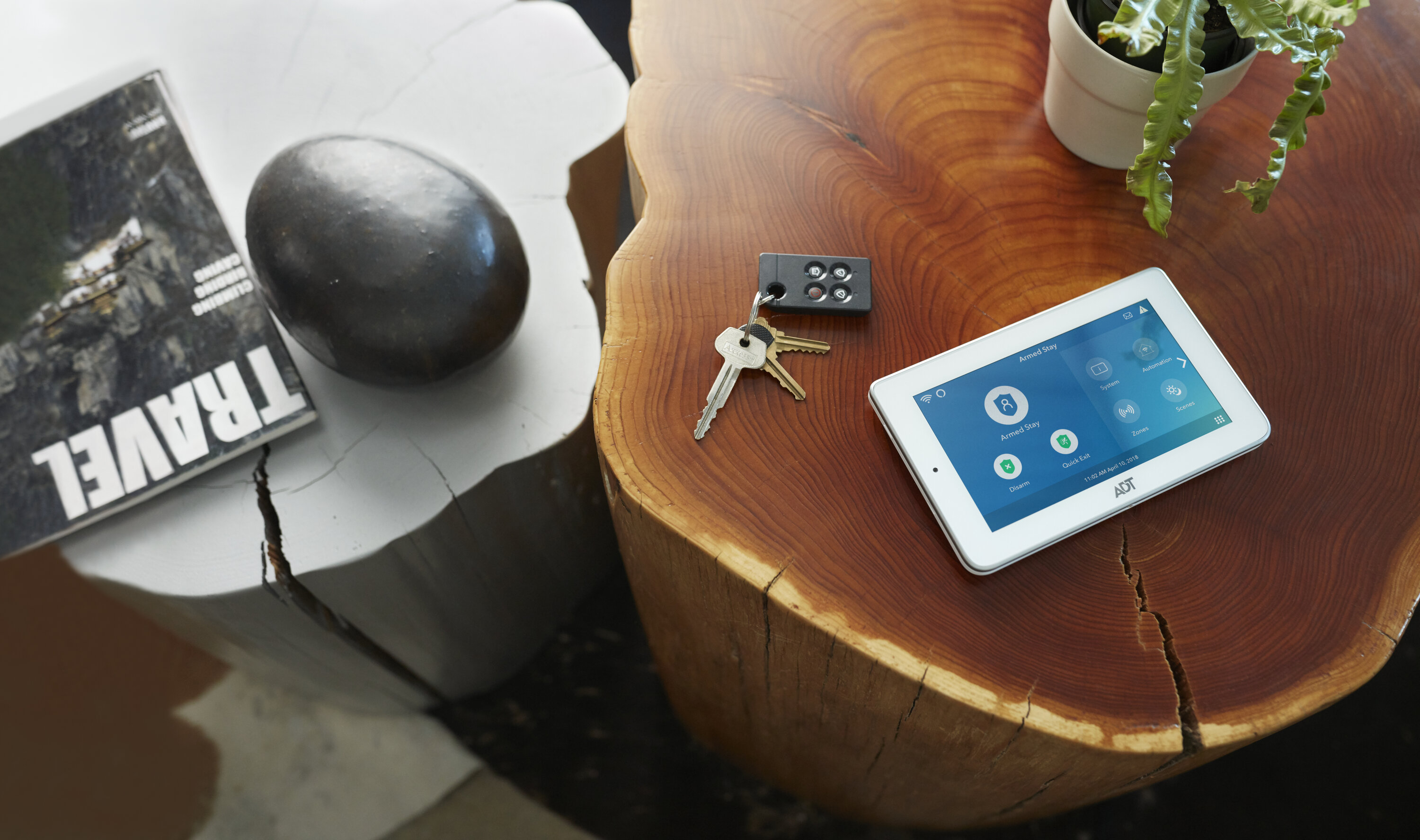 An ADT security touchscreen device sits on a wooden coffee table next to keys, a green plant, and a travel book