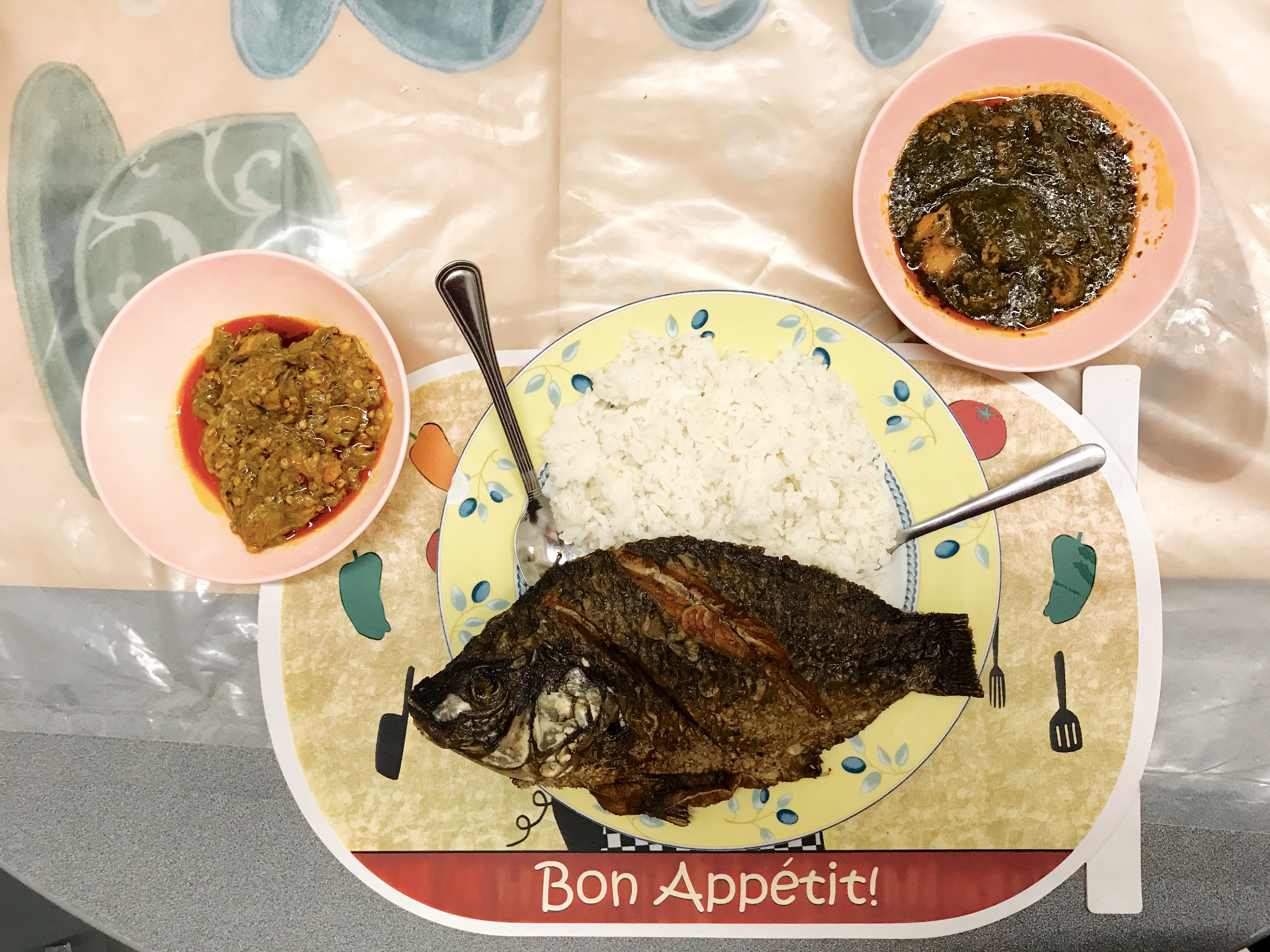 A whole fried tilapia sits on a plate on a pile of white rice. Underneath is a placemat that reads “Bon Appetit!” Two pink bowls hold side dishes.