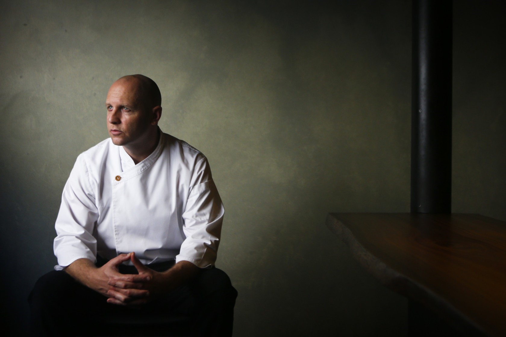 A chef looks out of a window while wearing whites in a semi-dark room.