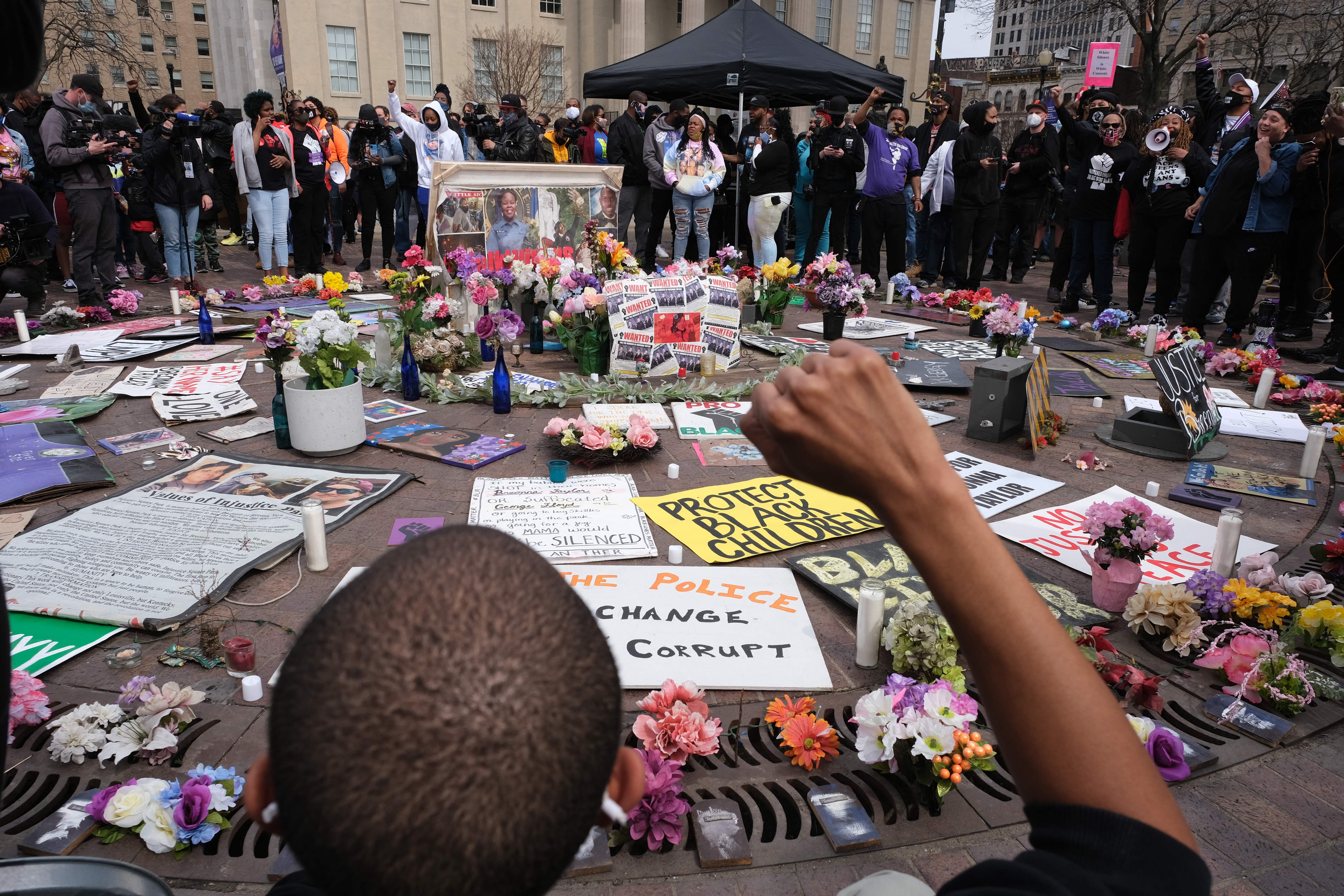 About 100 people stand in a circle; in the middle are signs with messages like “Protect Black Children” and “Change Corrupt Police” along with dozens of bouquets. In the foreground, a Black protester whose back is to the camera raises a fist.
