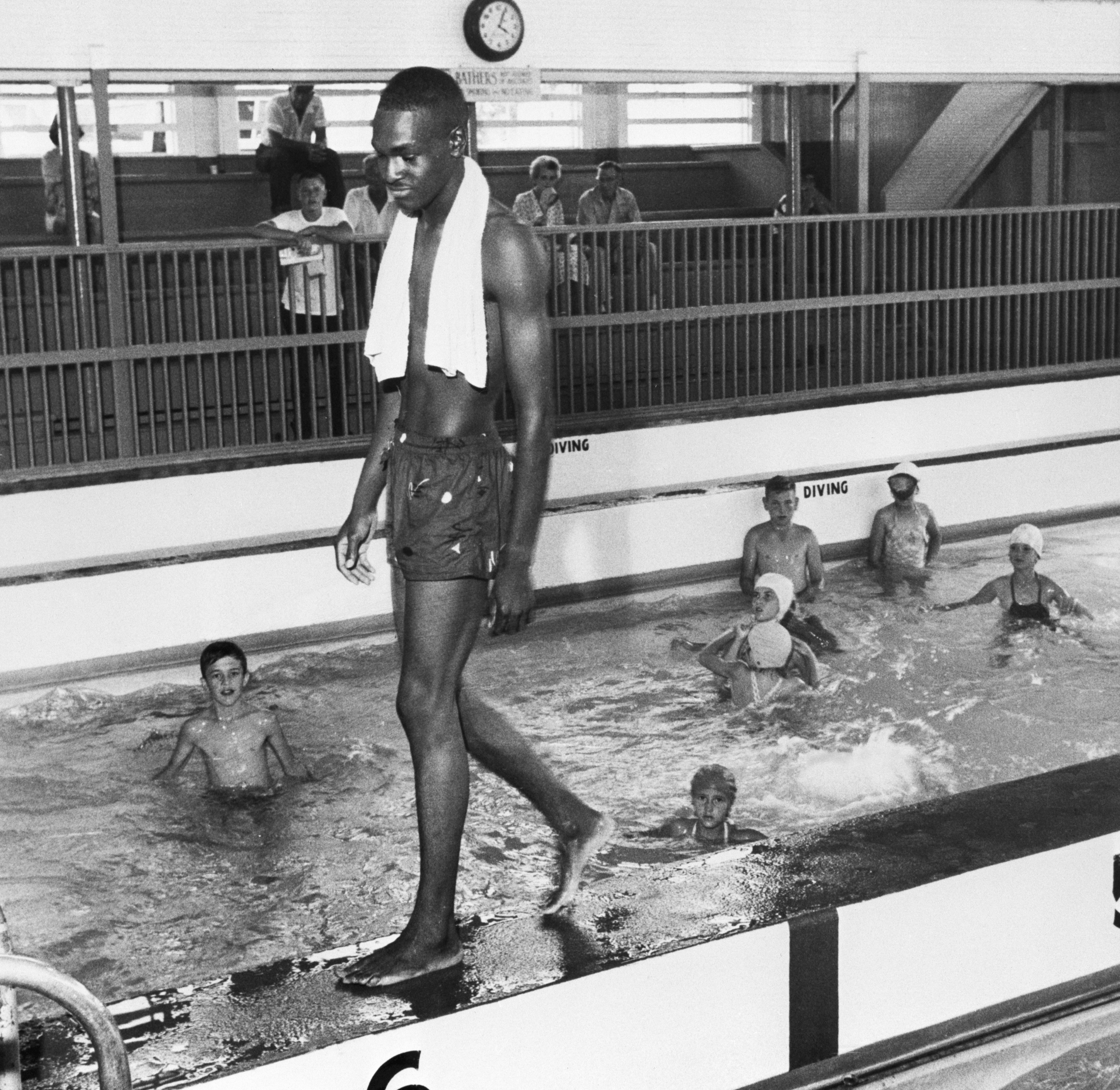 A black-and-white photo of a black man in swim trunks with a towel around his shoulders walking along the edge of an indoor pool.