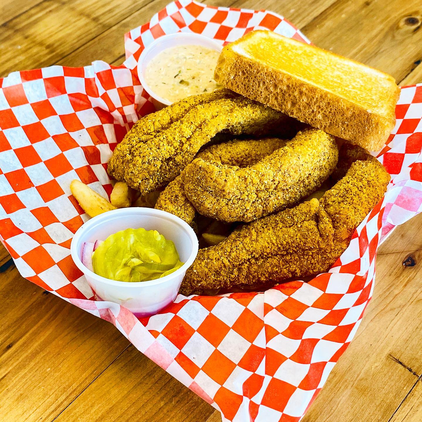 fried catfish tenders with thick toast, pickles, fries and tarter sauce in a plastic basket lines with red and white checkered paper