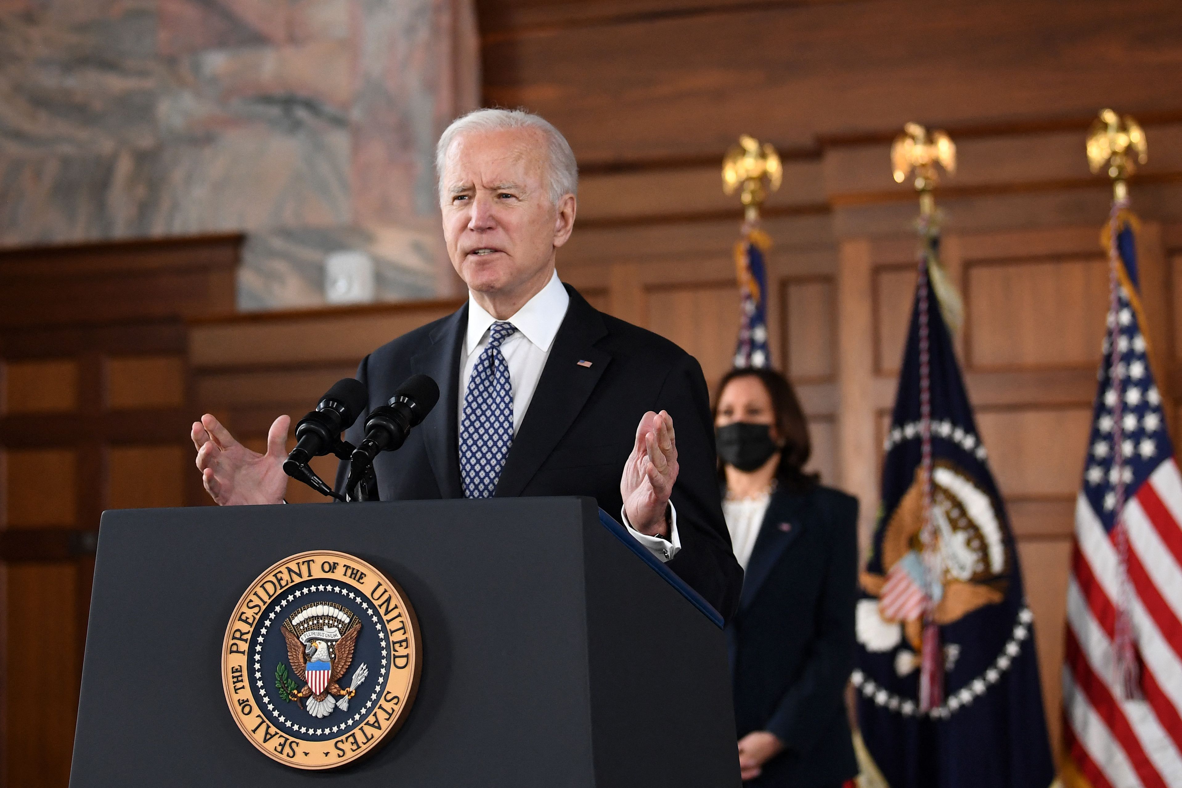 Biden, in a dark suit and blue tie, speaks at a podium bearing the seal of the US. Vice President Kamala Harris stands behind him, in a black suit and white blouse.