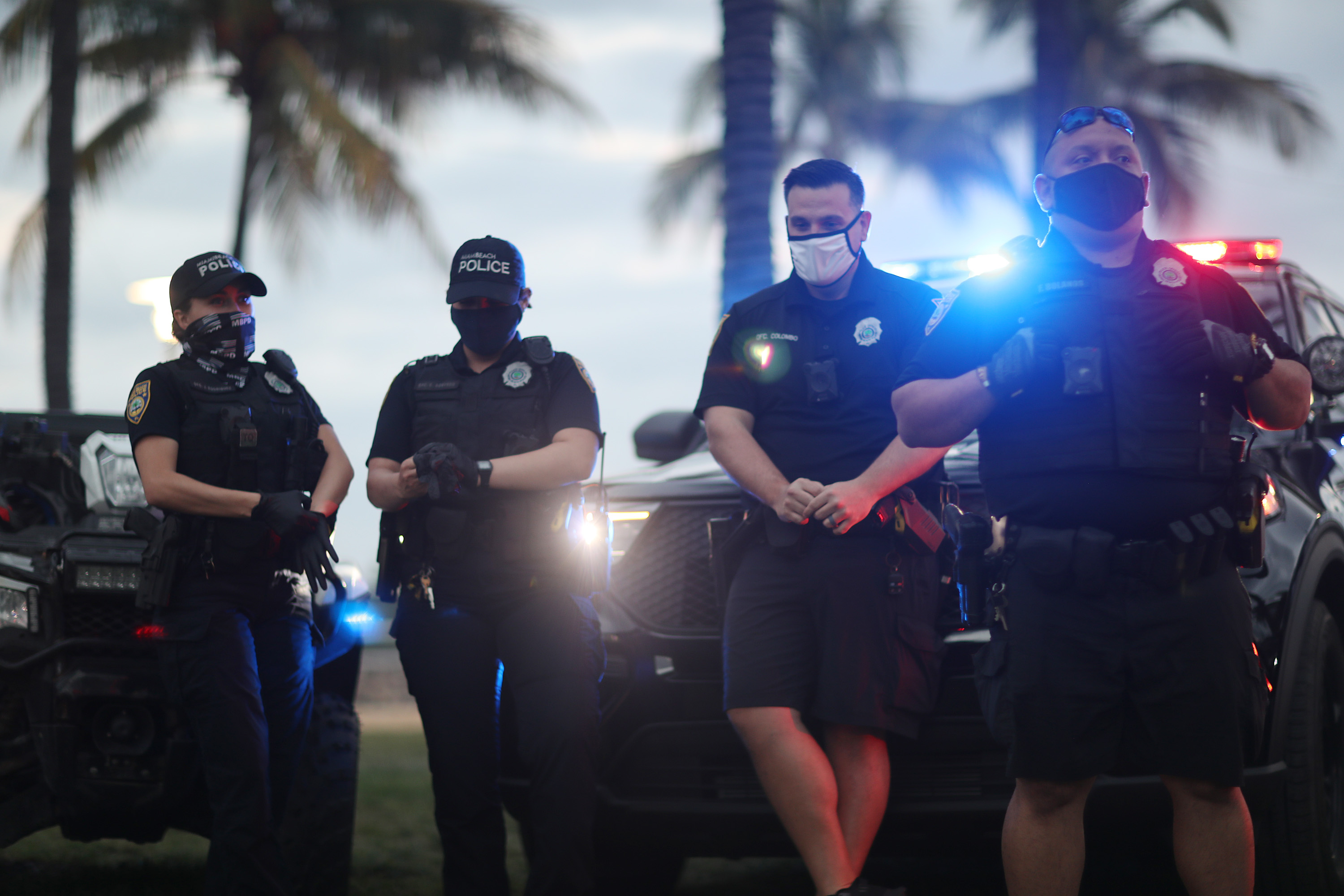 Four officers, two women and two men, stand in front of police cars with flashing lights. All are in navy blue uniforms, and all are wearing masks. 