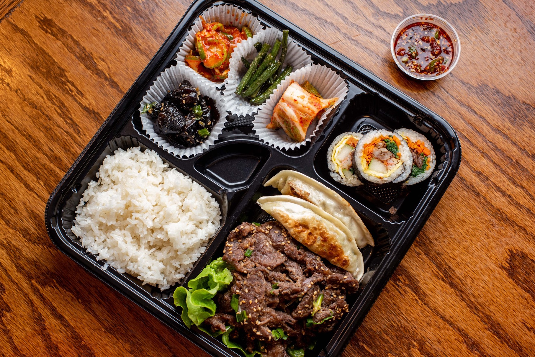 A black partitioned takeout container with white rice and beef bulgogi as main entrees alongside dumpling and banchan from Woo Nam Jeong Stone Bowl House in Doraville