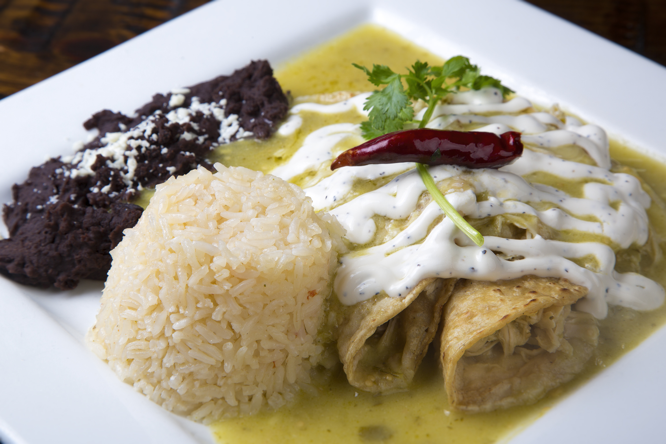 Two Enchiladas verde sit atop a white square plate drizzled with chipotle cream and verde sauce. A cylindrical-shaped stack of Mexican rice sits beside the enchiladas with a dollop of refried beans behind it.