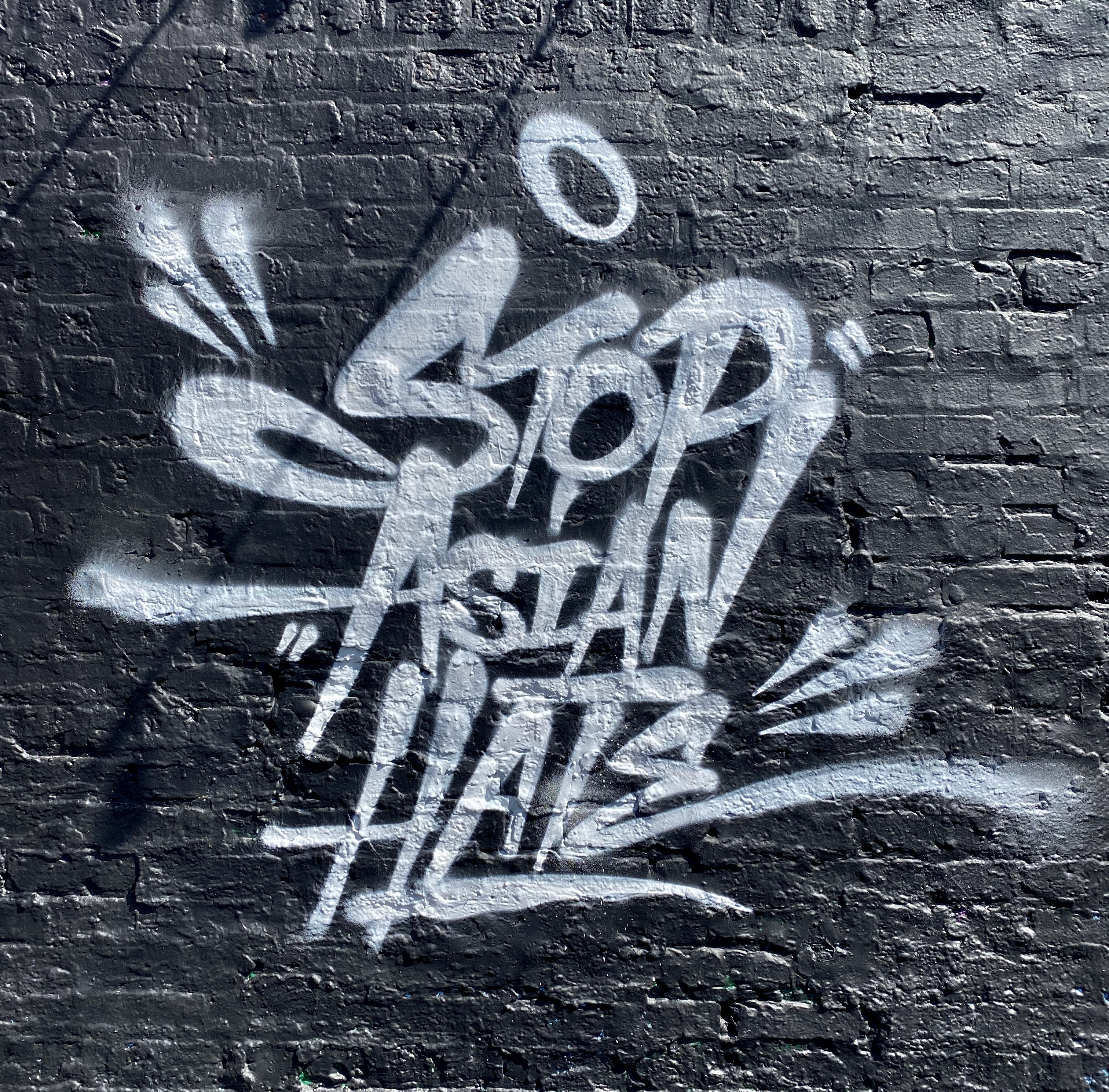 A graffiti tag reading “stop Asian hate” using white paint on a black brick wall.