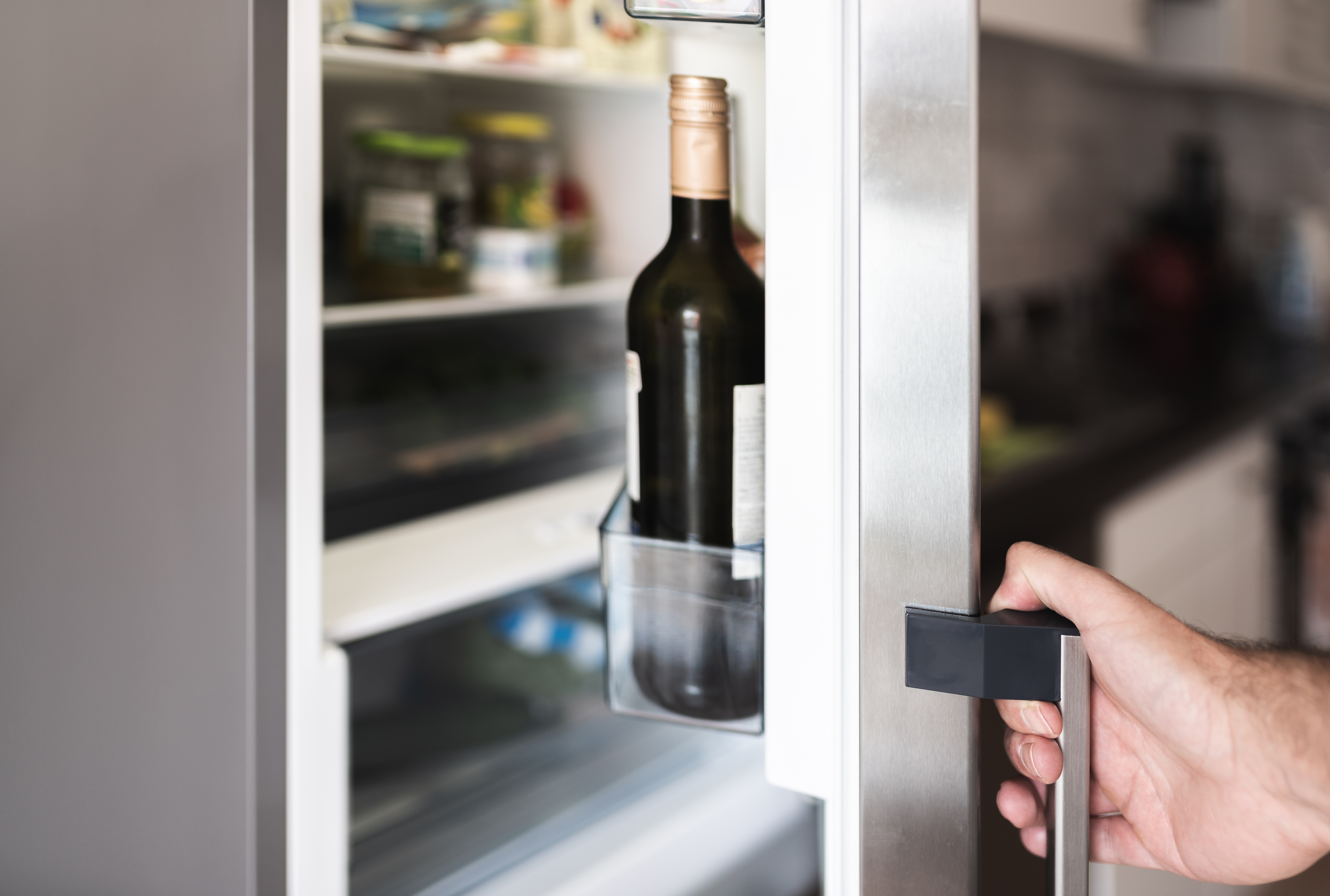 Sleek refrigerator door being opened by a white male hang, with a bottle of wine visible on the door shelf