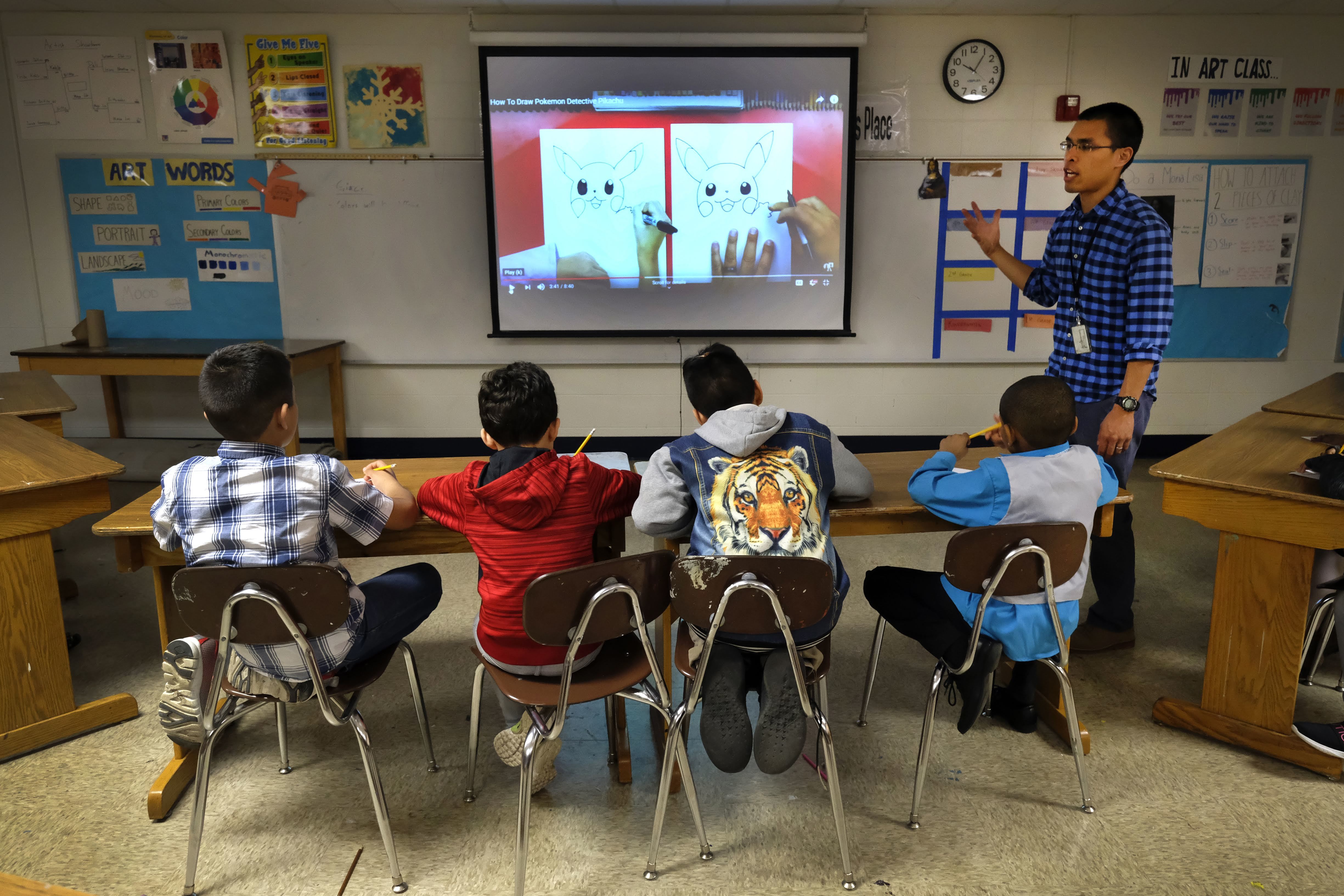 An April 2019 photo shows a teacher next to a smart board talking to four male students sitting at a table and looking at the board.