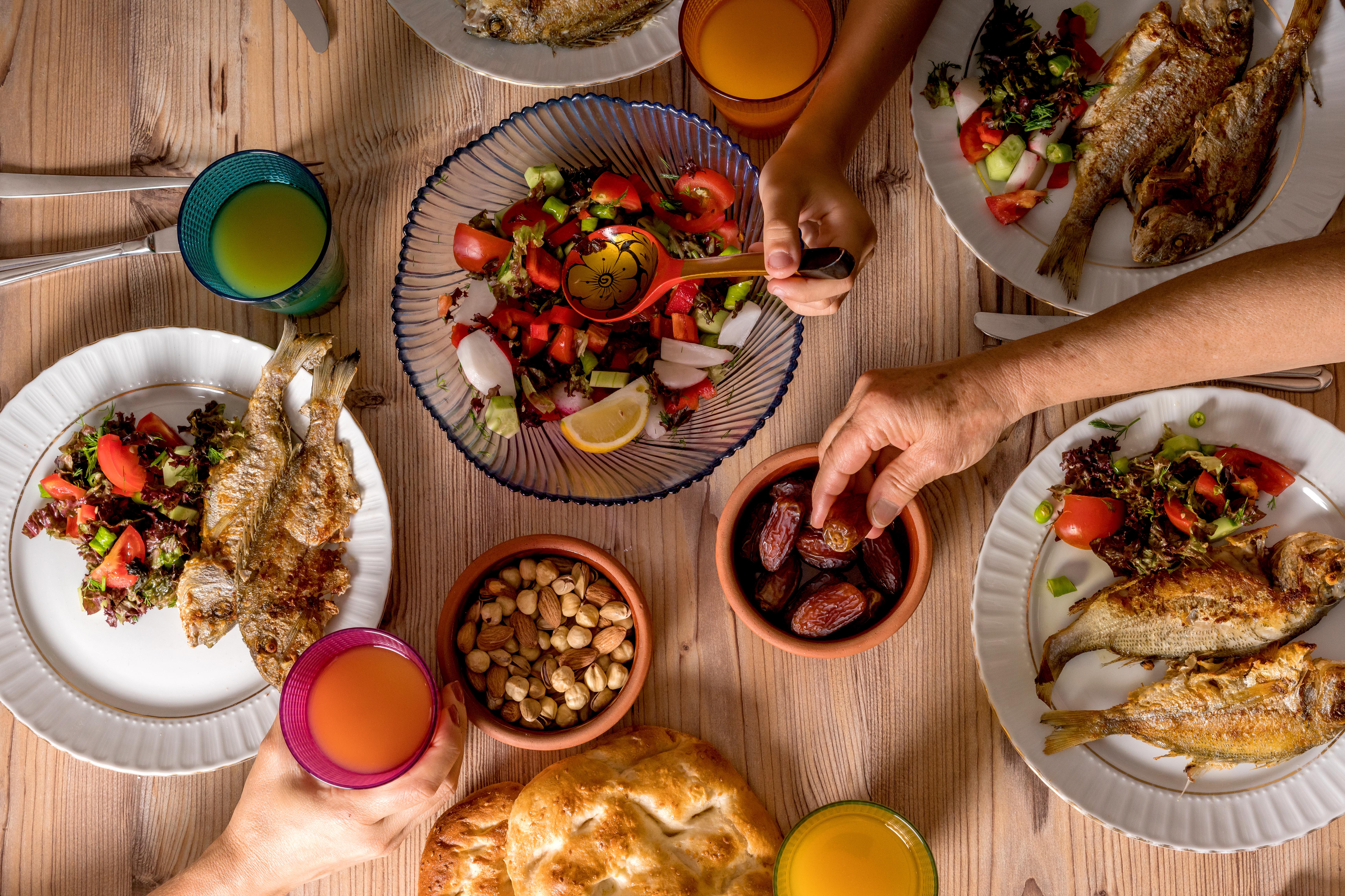 An overhead shot of an Iftar meal on a wooden dining table. Hands reach in from the top, right, and bottom to grab dates, mix a salad, and hold a cup.