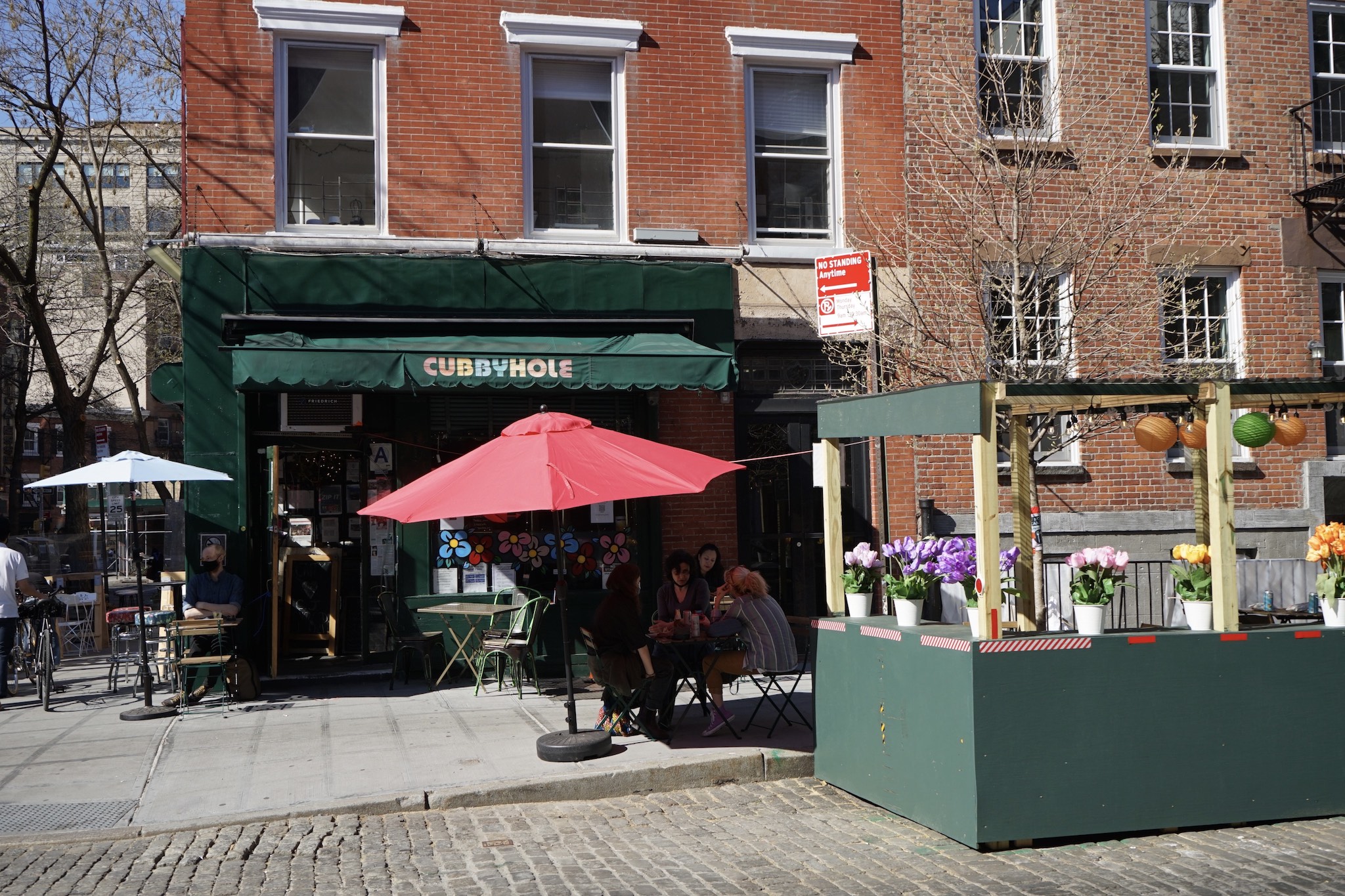 A green-painted exterior of a bar with red and white umbrellas unfurled over tables outside. A green plywood patio sits on the cobblestone street next to the bar entrance.