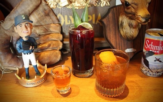 A Seattle Mariners bobblehead doll sits to the left, in front of a vintage baseball mitt; next to that are three cocktails and a can of Schmidt beer; behind the can of beer is a mounted deer’s head.