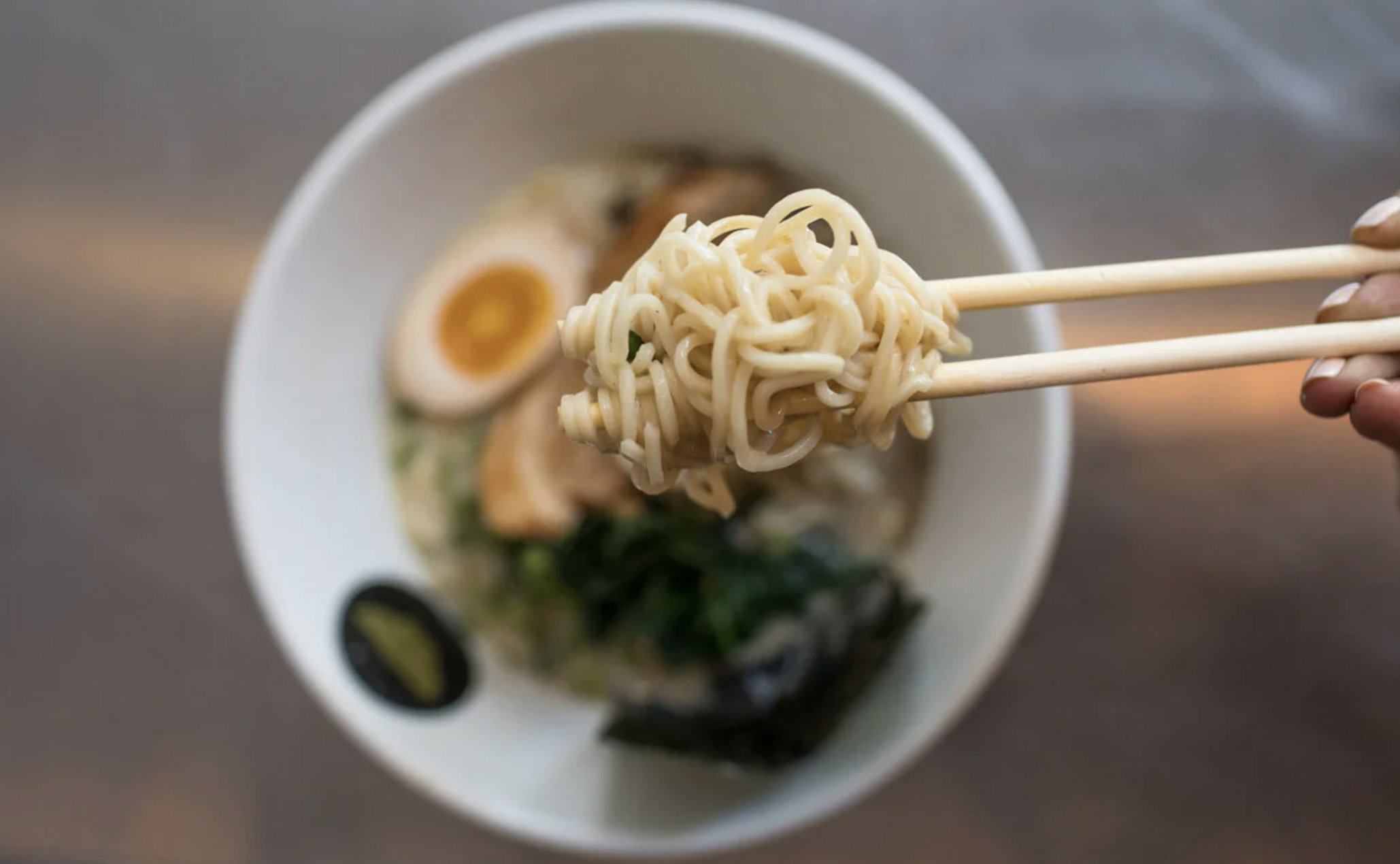 Chopsticks lift a tangle of ramen noodles from a bowl filled with hard-boiled egg, seaweed, and broth; topdown view.