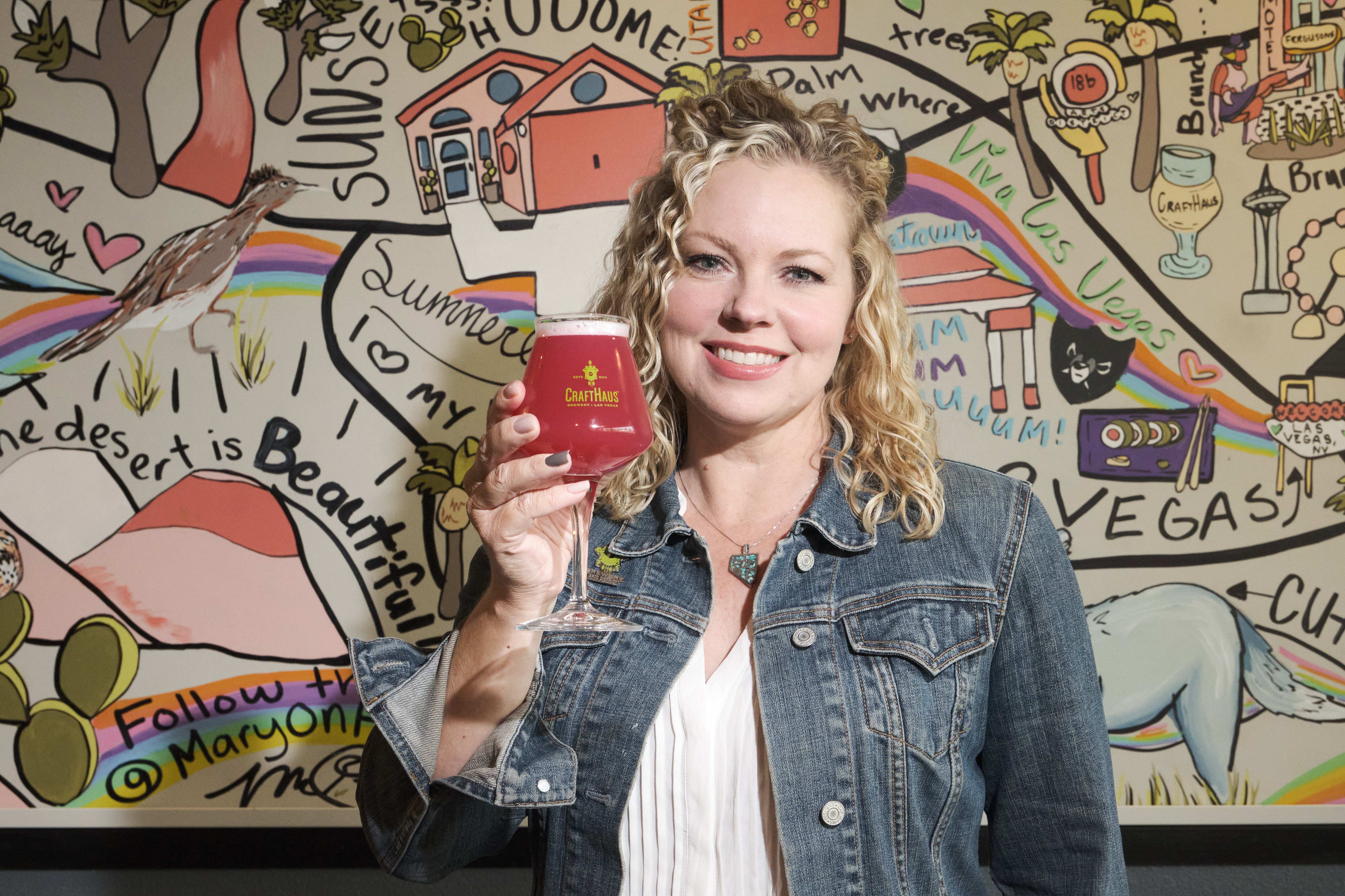A woman stands in front of a mural holding a beer