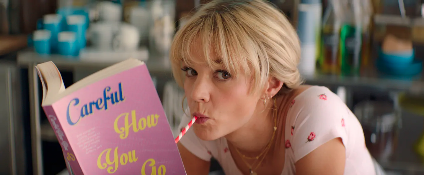 A young woman in pigtails drinks from a milkshake and reads a pink book entitled “Careful How You Go.”