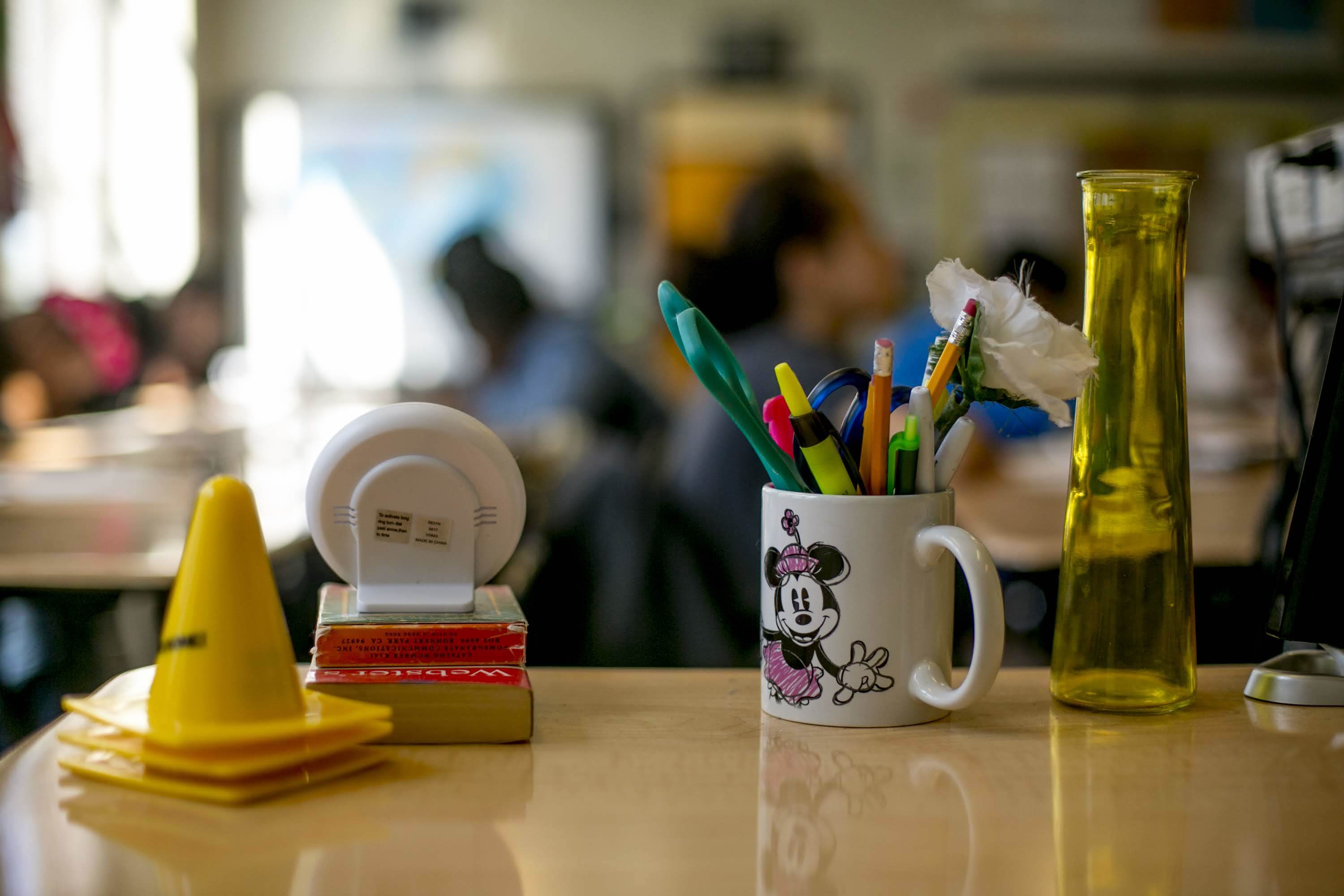Markers, pencils and pens in a Minnie Mouse coffee cup, small yellow cones, a dictionary, and other items sit atop a desk in a classroom.