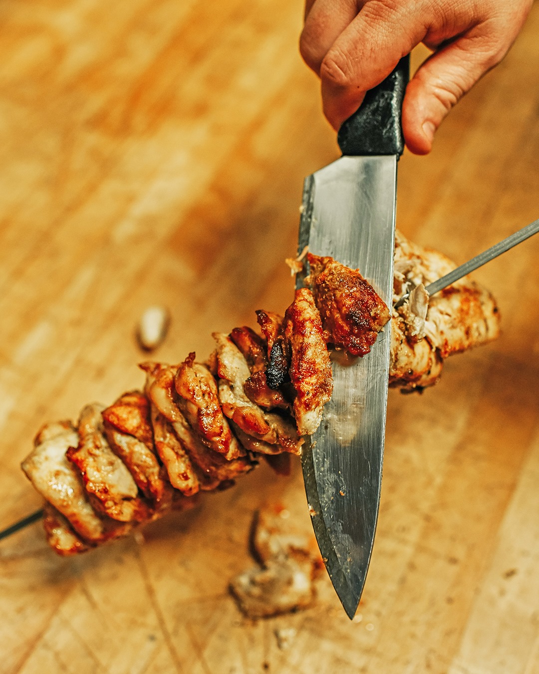 A sharp knife slices pieces of spit-fire roasted chicken from a metal skewer
