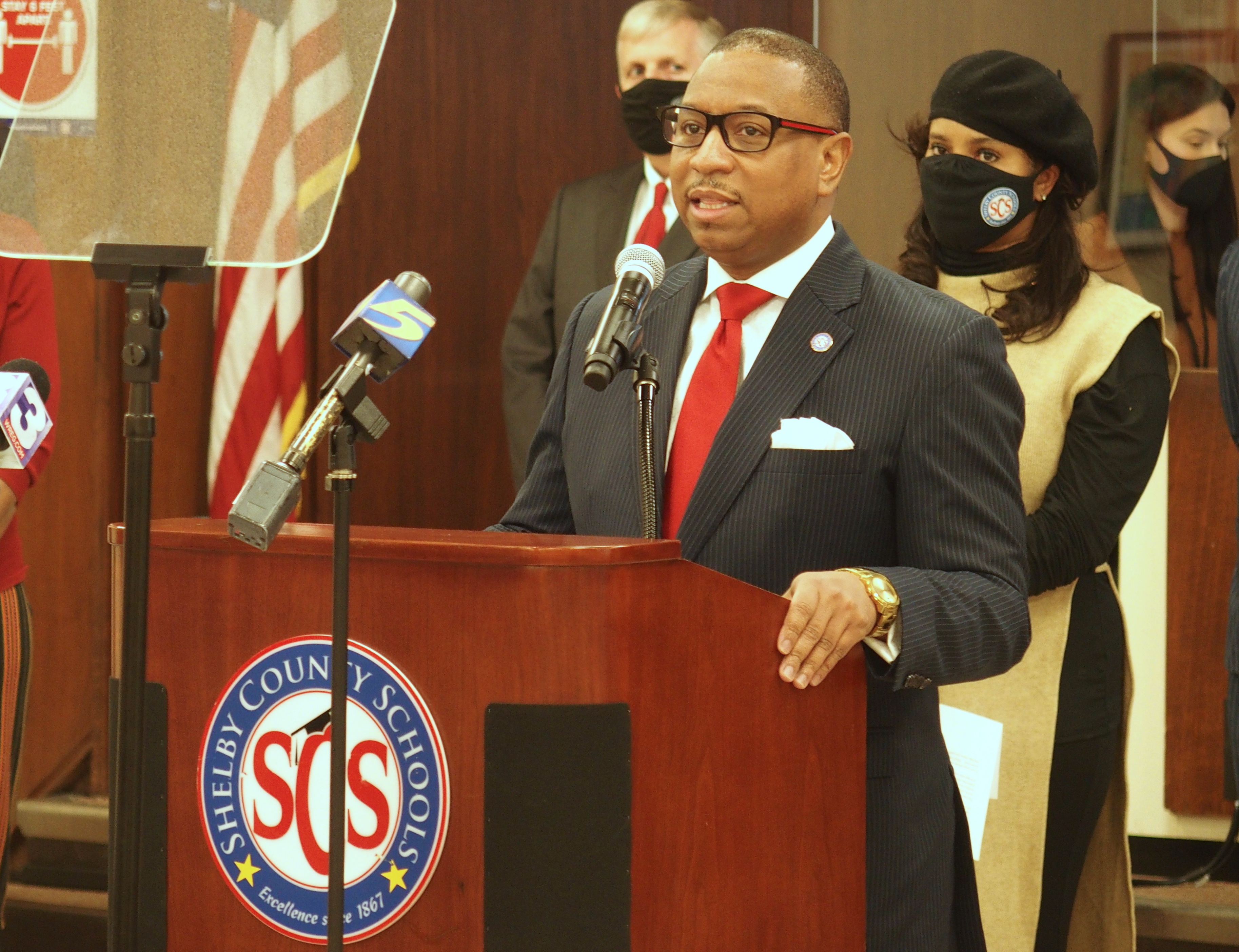 Superintendent Joris Ray stands at a podium with microphones during a press conference as school board chairwoman Miska Clay Bibbs, wearing a mask, looks on from behind.